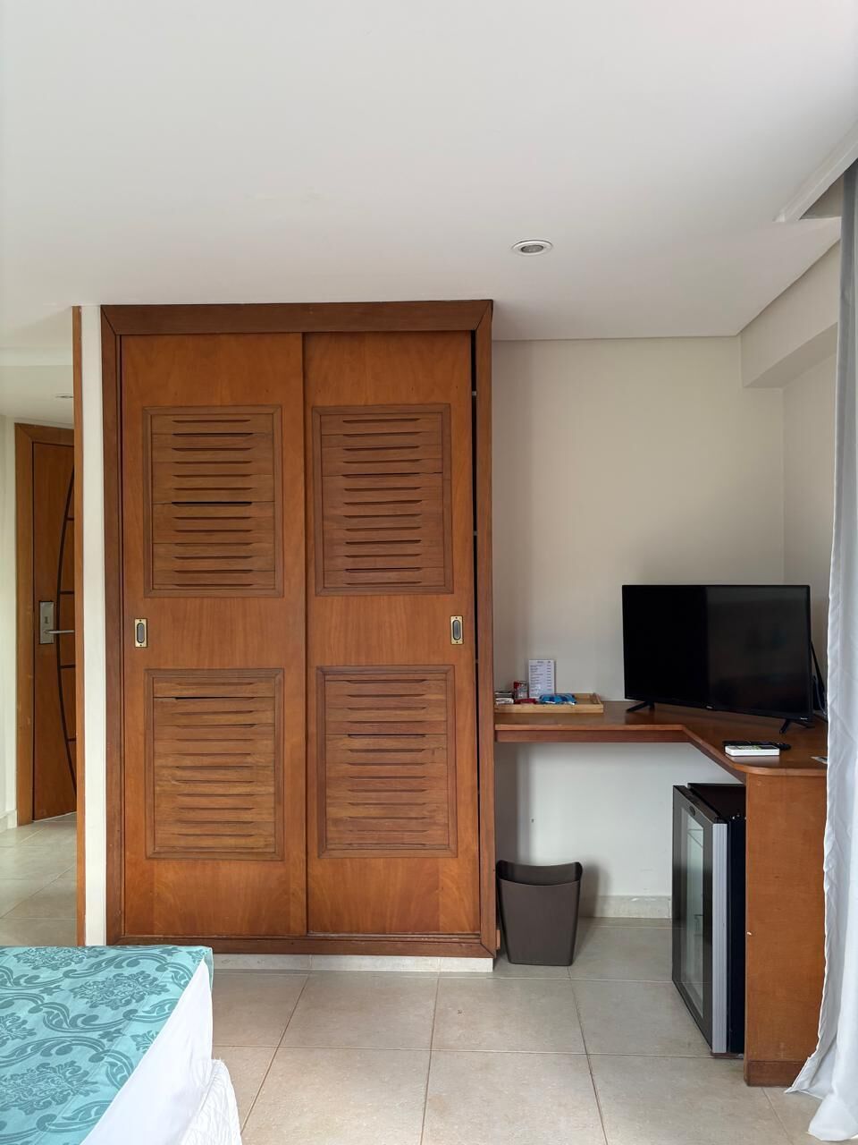 A room with a wooden wardrobe, a television, a small refrigerator, and a trash bin.