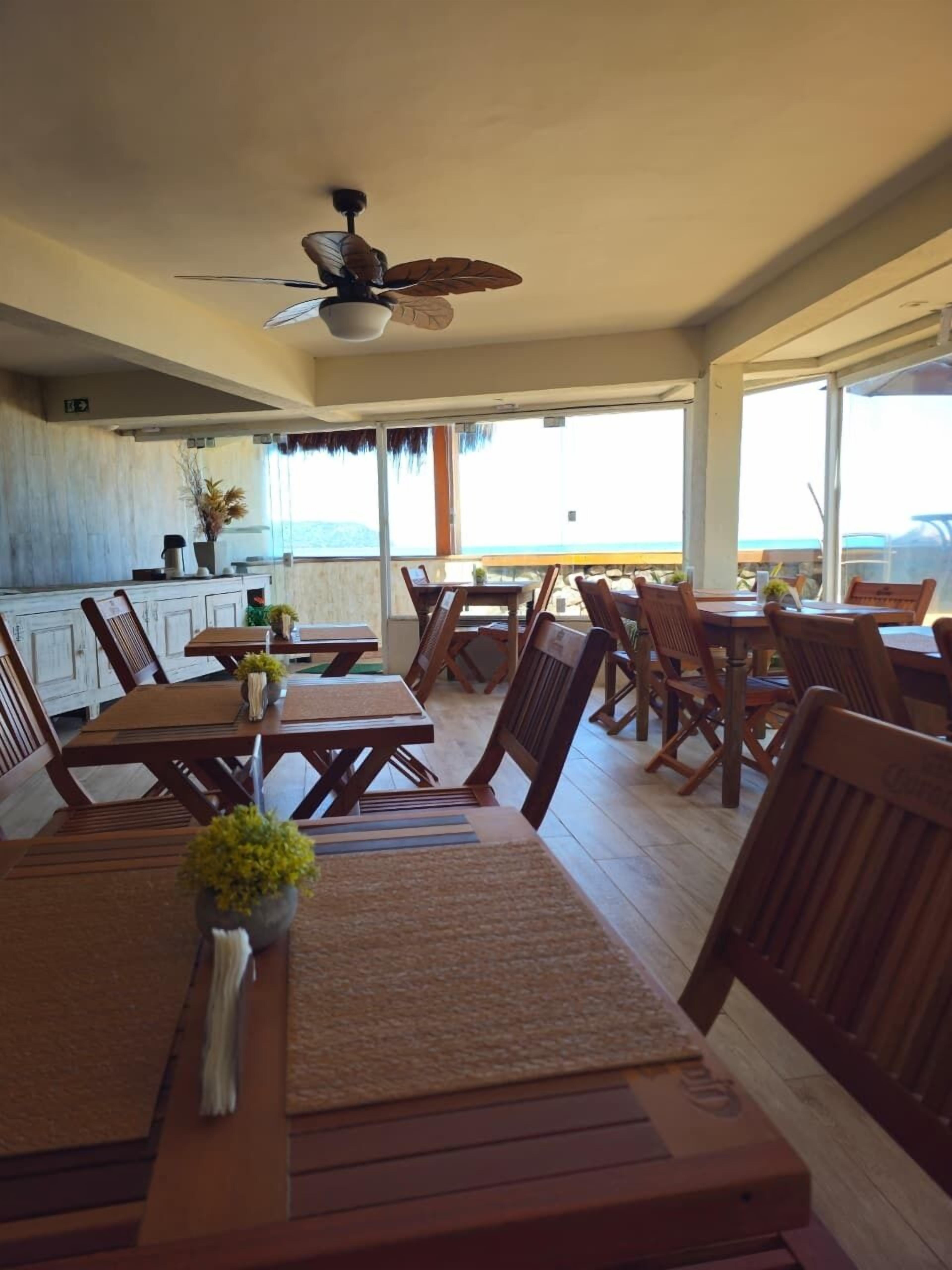 A dining area with wooden tables and chairs, a ceiling fan, and a view of the ocean.