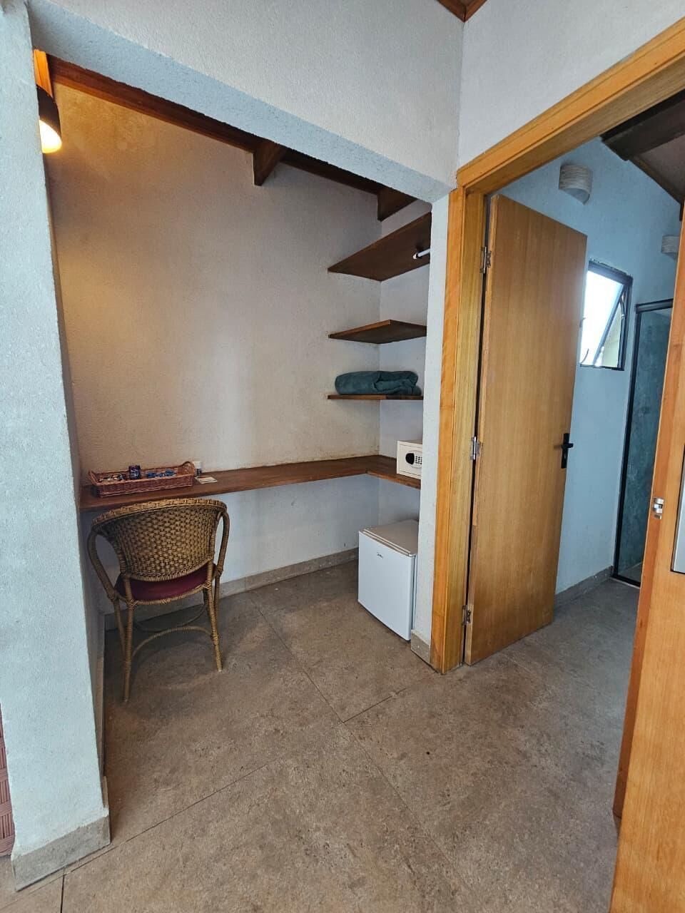 A compact room with a small desk, a chair, a refrigerator, and a wooden door leading to another room.