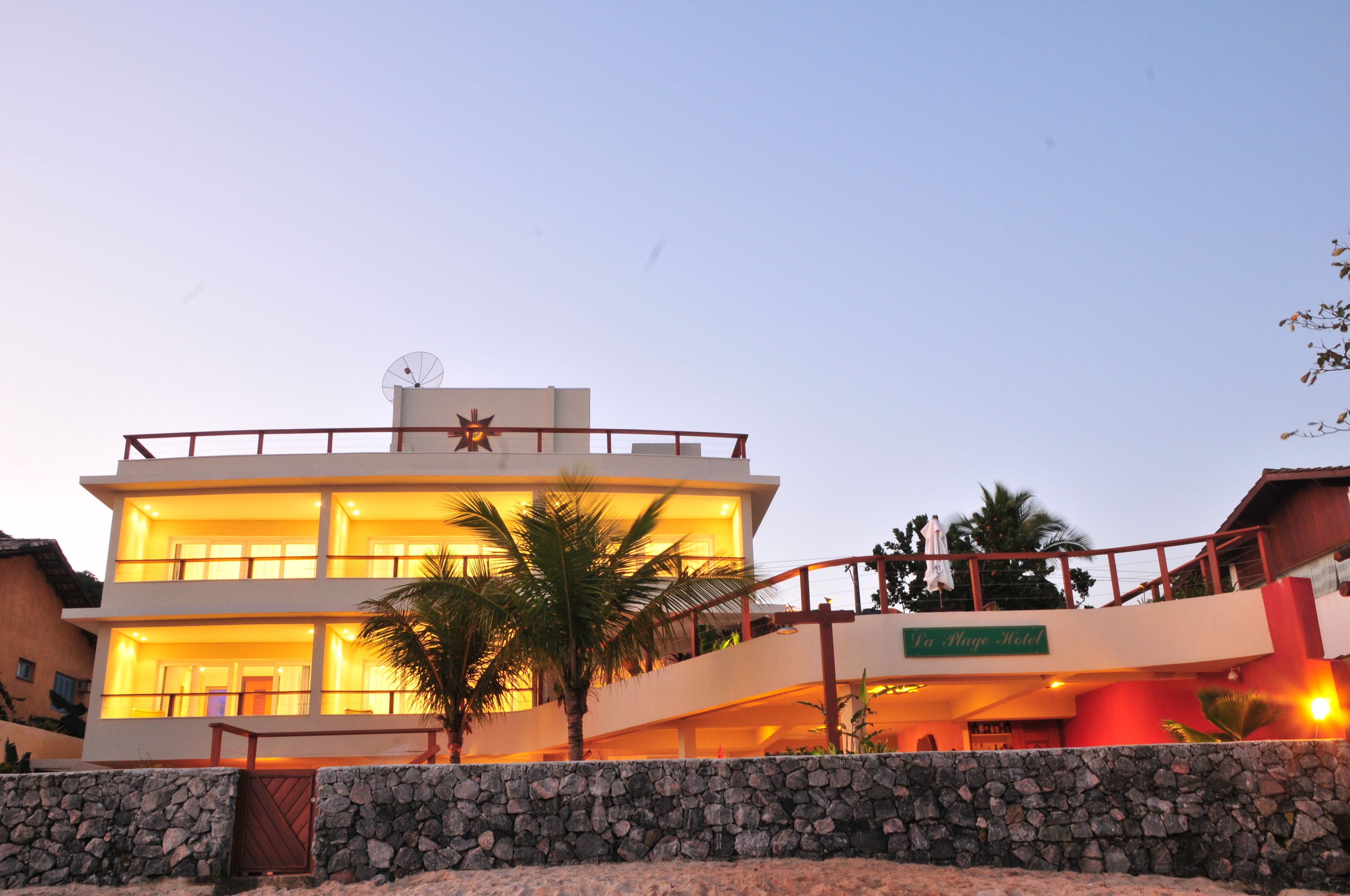 Un hotel de dos pisos con balcón, un muro de piedra en primer plano y una palmera.
