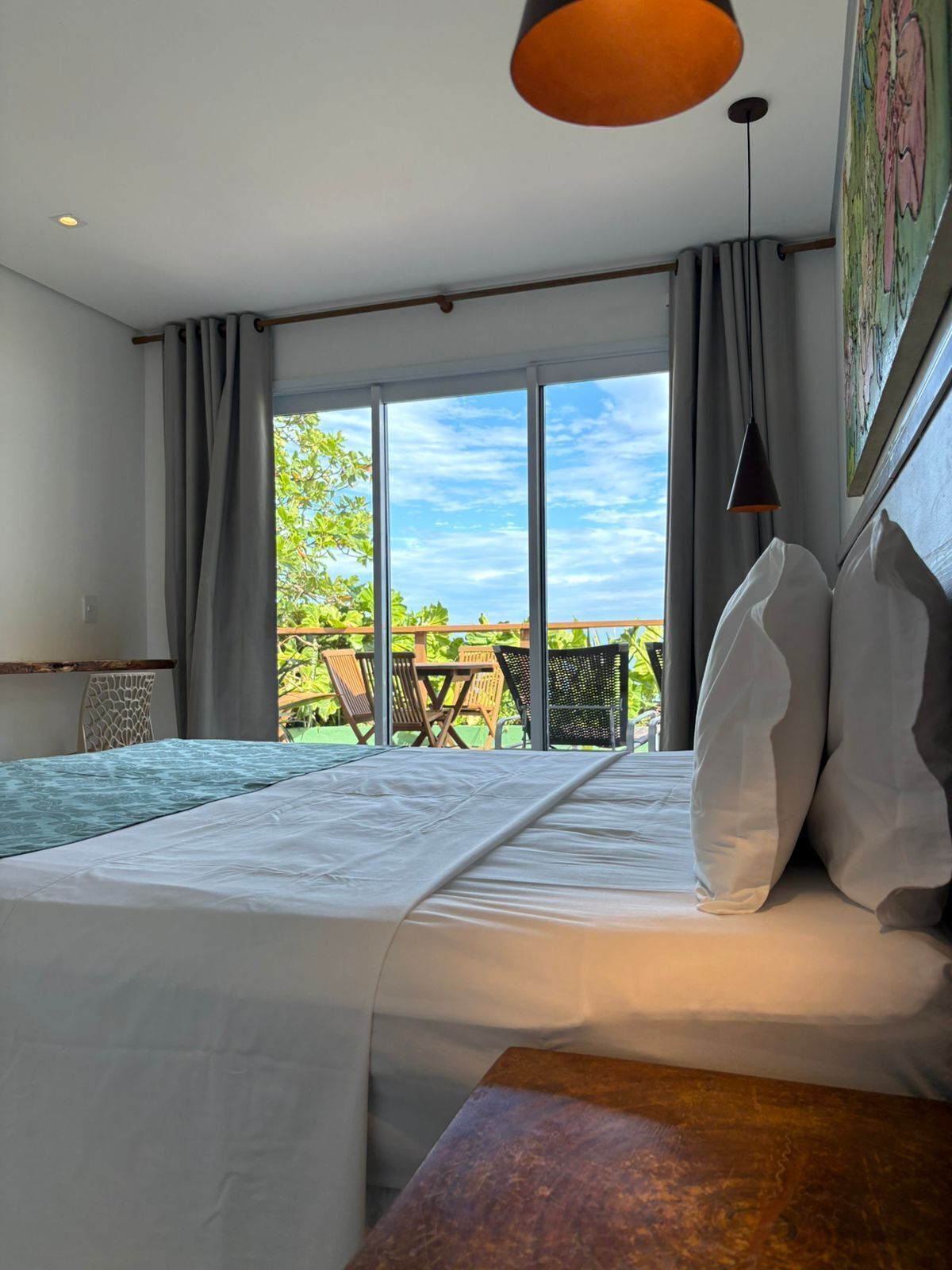 A bedroom with a large bed, a wooden nightstand, a hanging lamp, and a view of the outdoors.