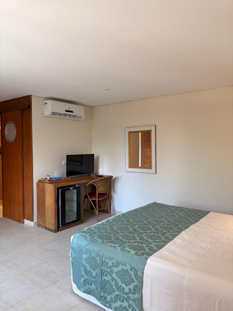 A hotel room with a bed, a desk with a television, a small refrigerator, and an air conditioning unit.