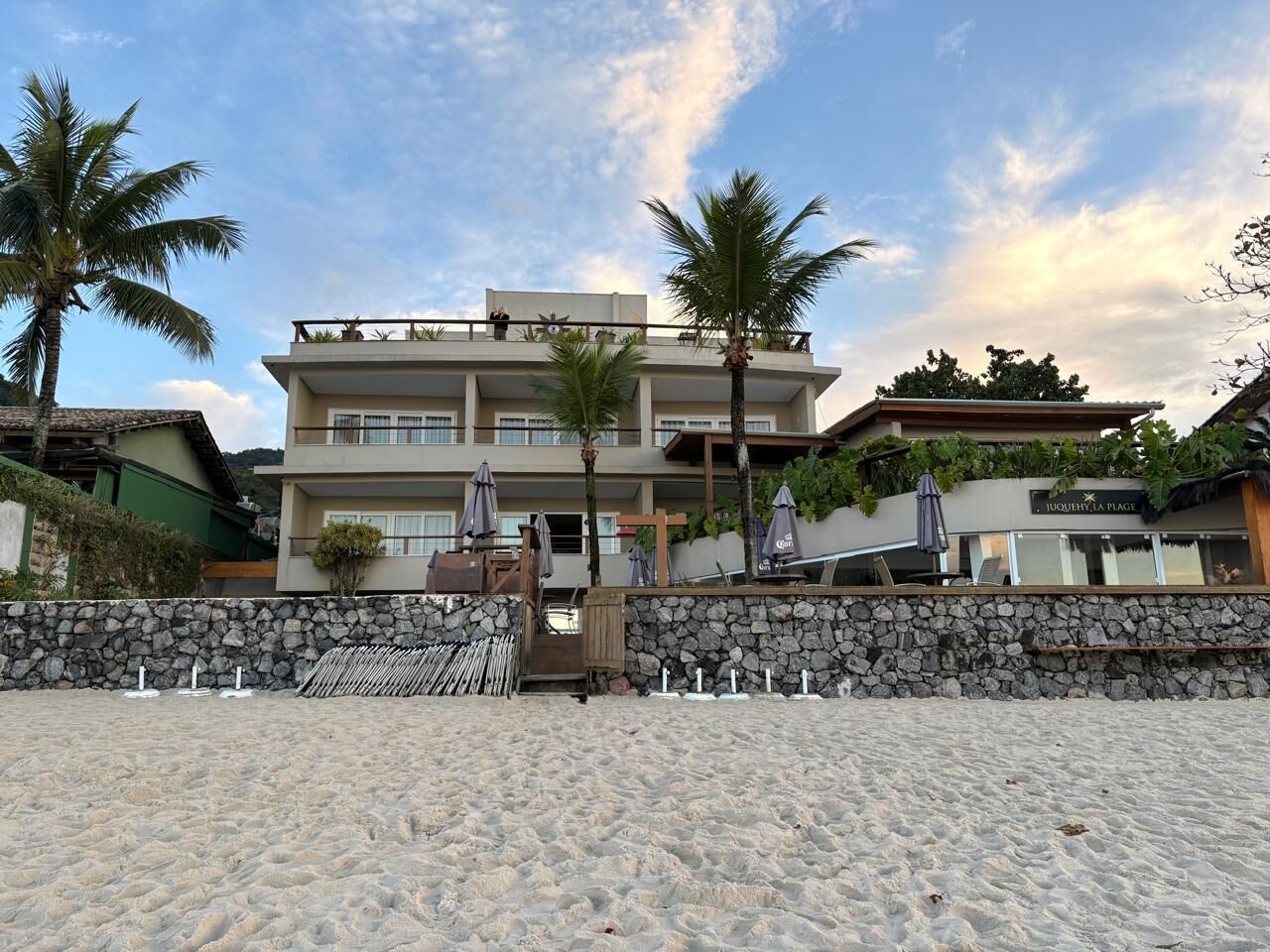 A beachfront hotel with a stone wall, palm trees, and a sign reading 'JUQUEYLA PLAGE'.