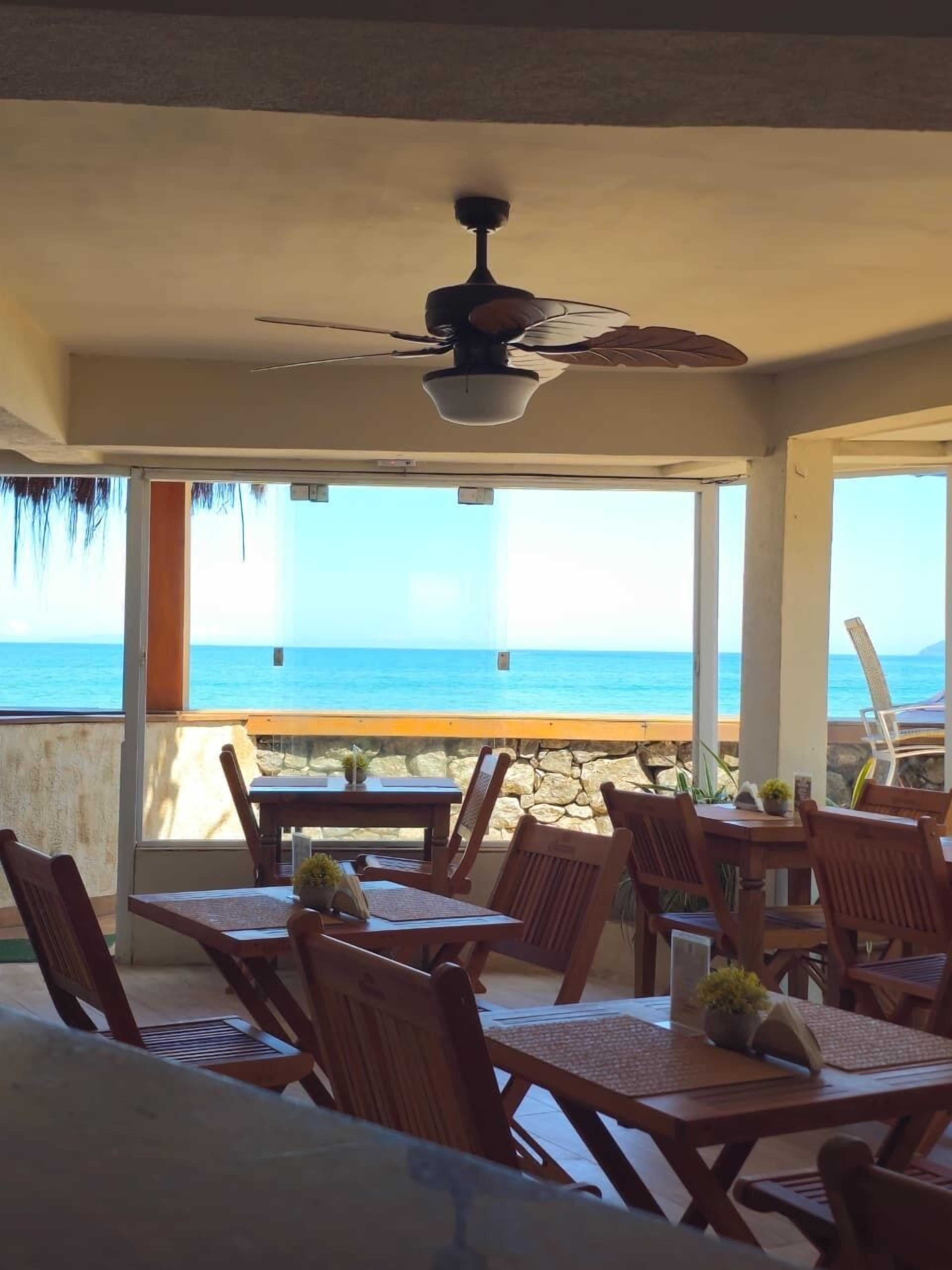 A covered outdoor seating area with a ceiling fan, wooden tables and chairs, and a view of the ocean.