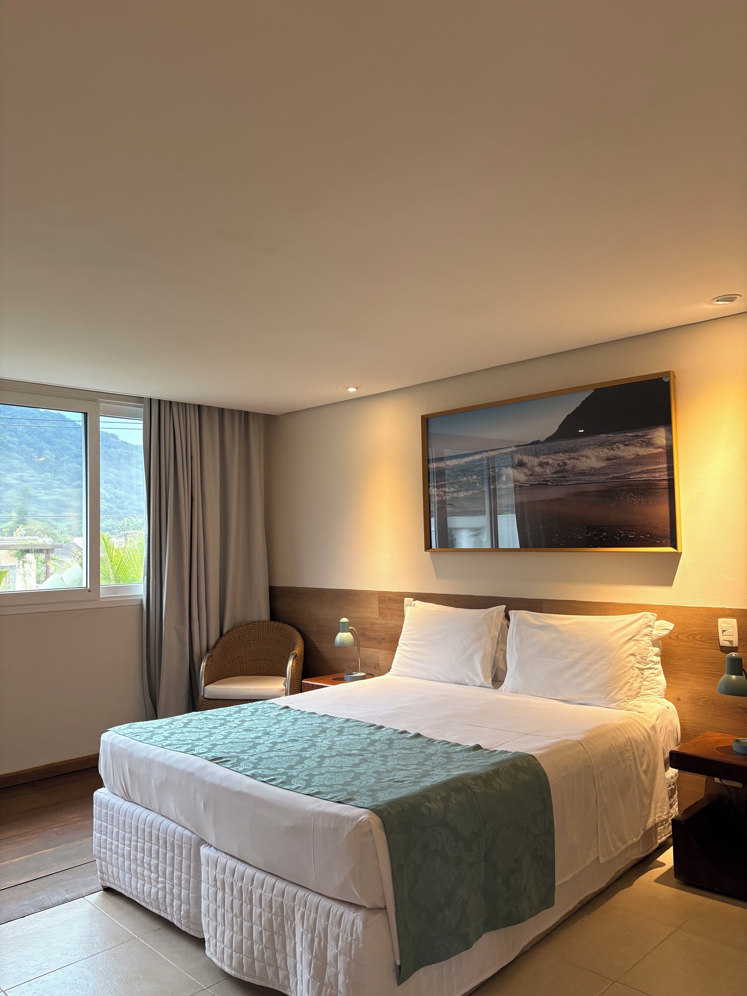 A hotel room with a bed, a chair, a nightstand, a lamp, a painting, and a window with curtains.