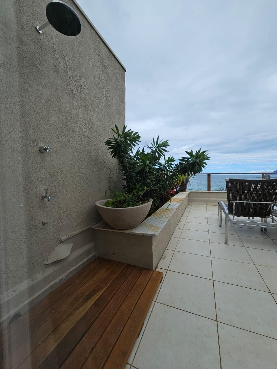 A balcony with a concrete wall, a large planter with plants, a wooden bench, and a view of the sea.