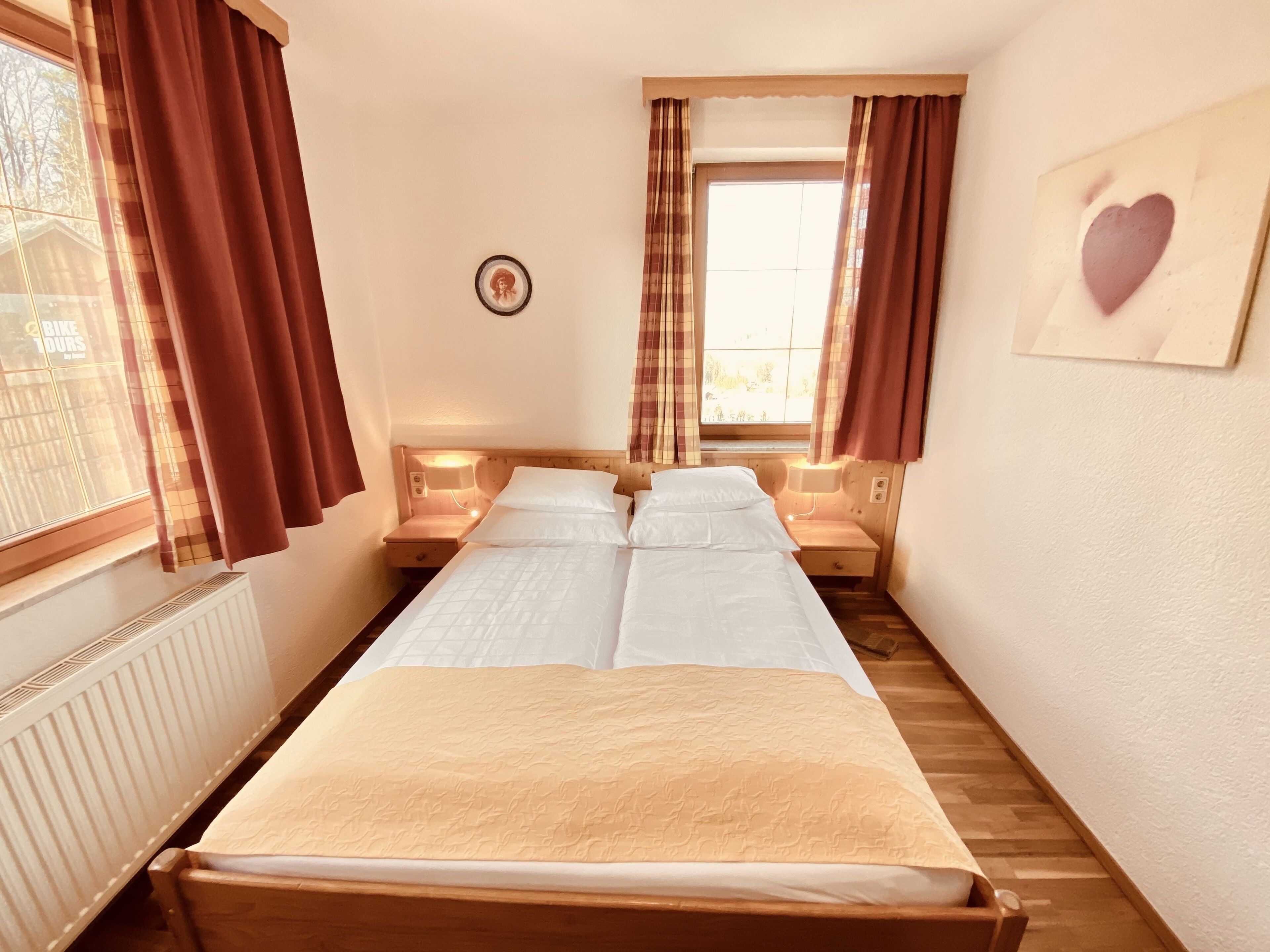 Una habitación pequeña y acogedora con una cama, dos mesitas de noche, un ventanal con cortinas, un radiador y un cuadro enmarcado en la pared.
