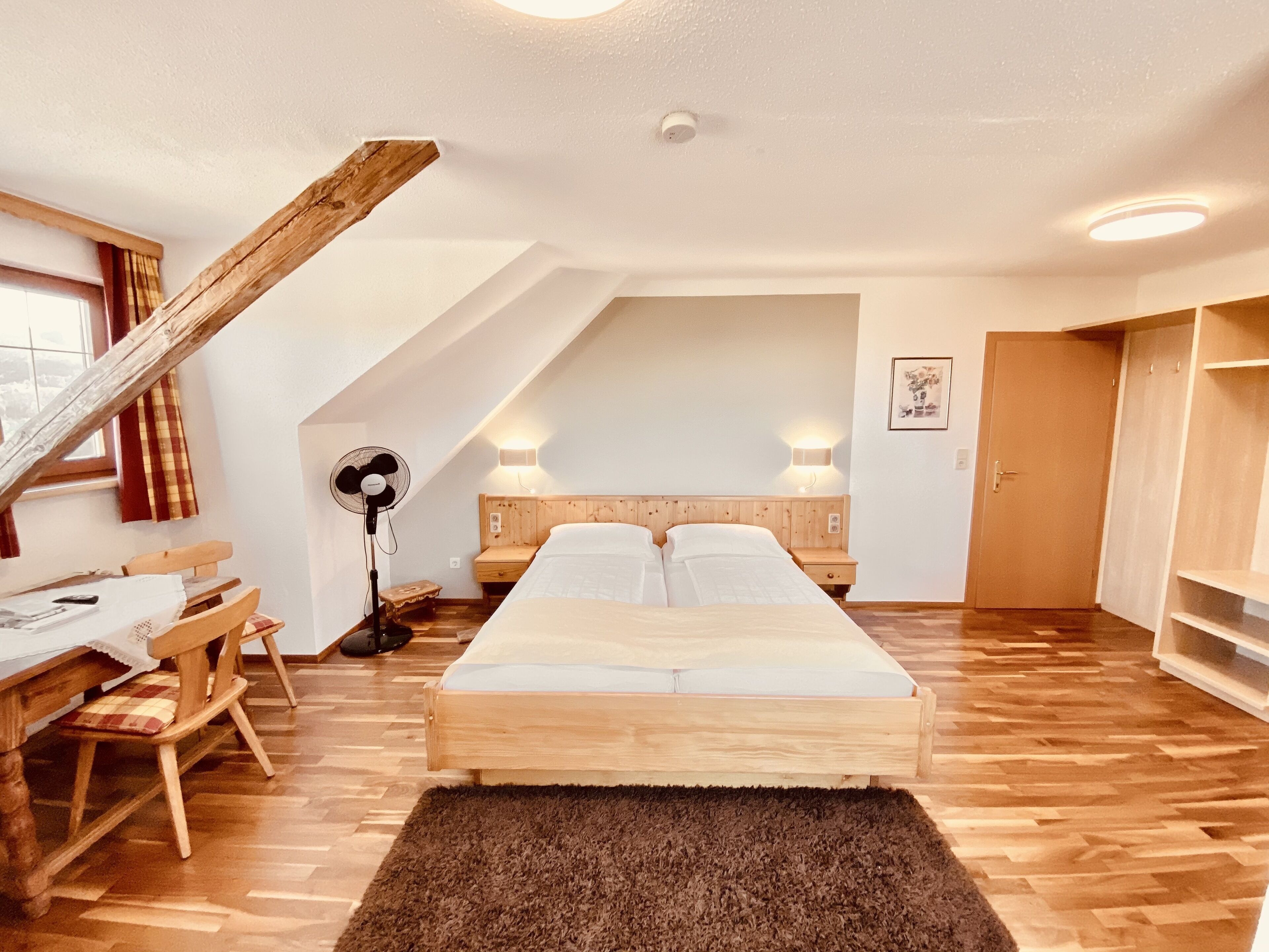 Un dormitorio con cama de madera, un escritorio, una silla, un ventilador de pie y techo inclinado.
