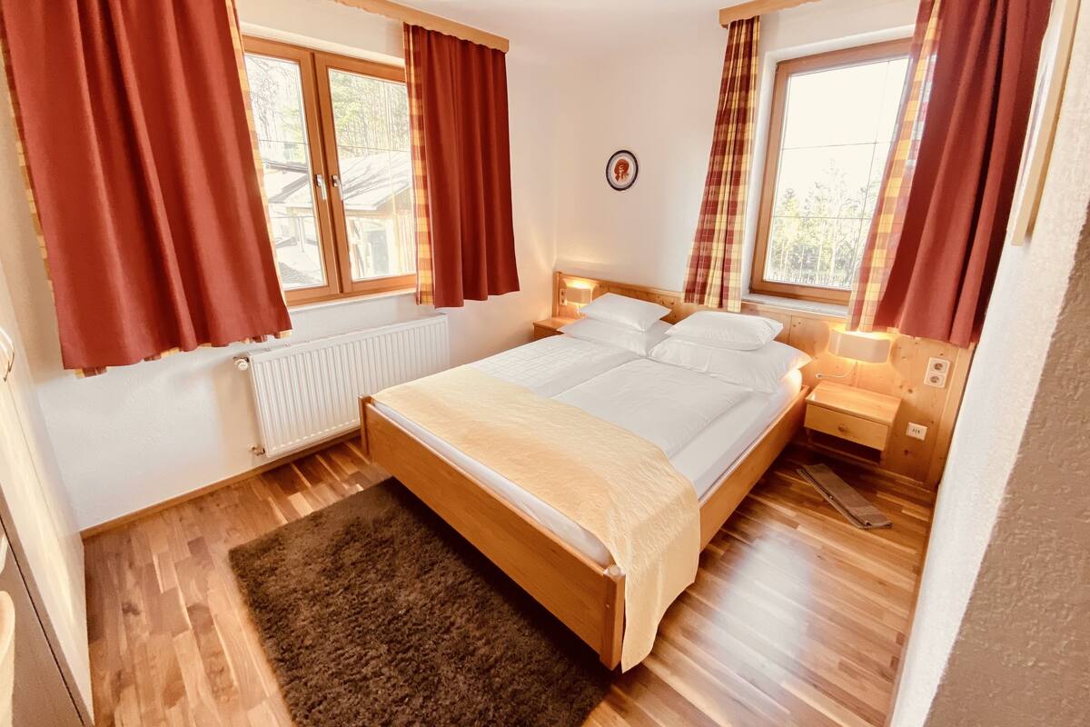 A double bed with a wooden frame, two bedside tables, a radiator, and large windows with curtains.