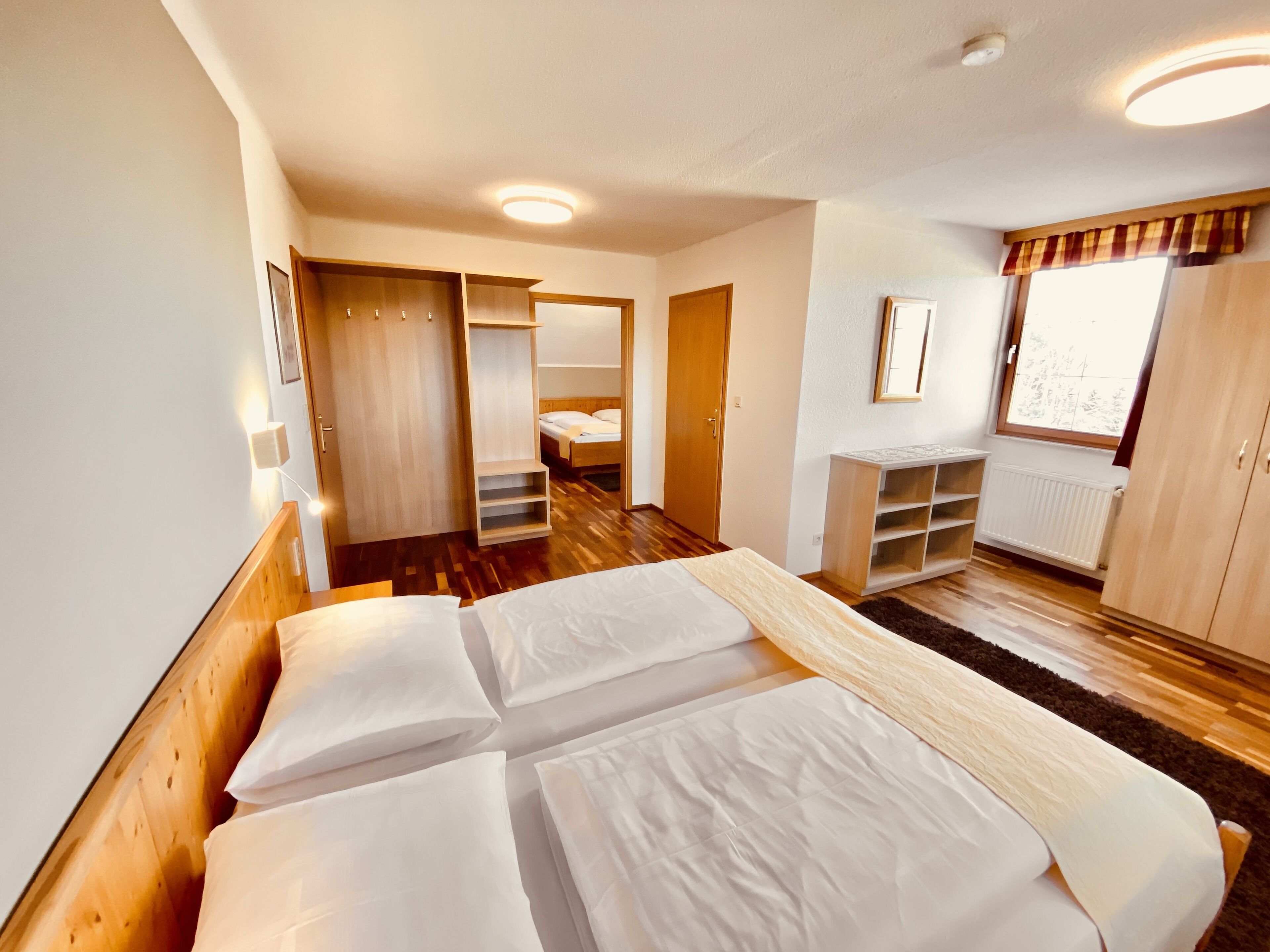 A bedroom with a large bed, wooden wardrobe, built-in shelving, a window with curtains, and a radiator.