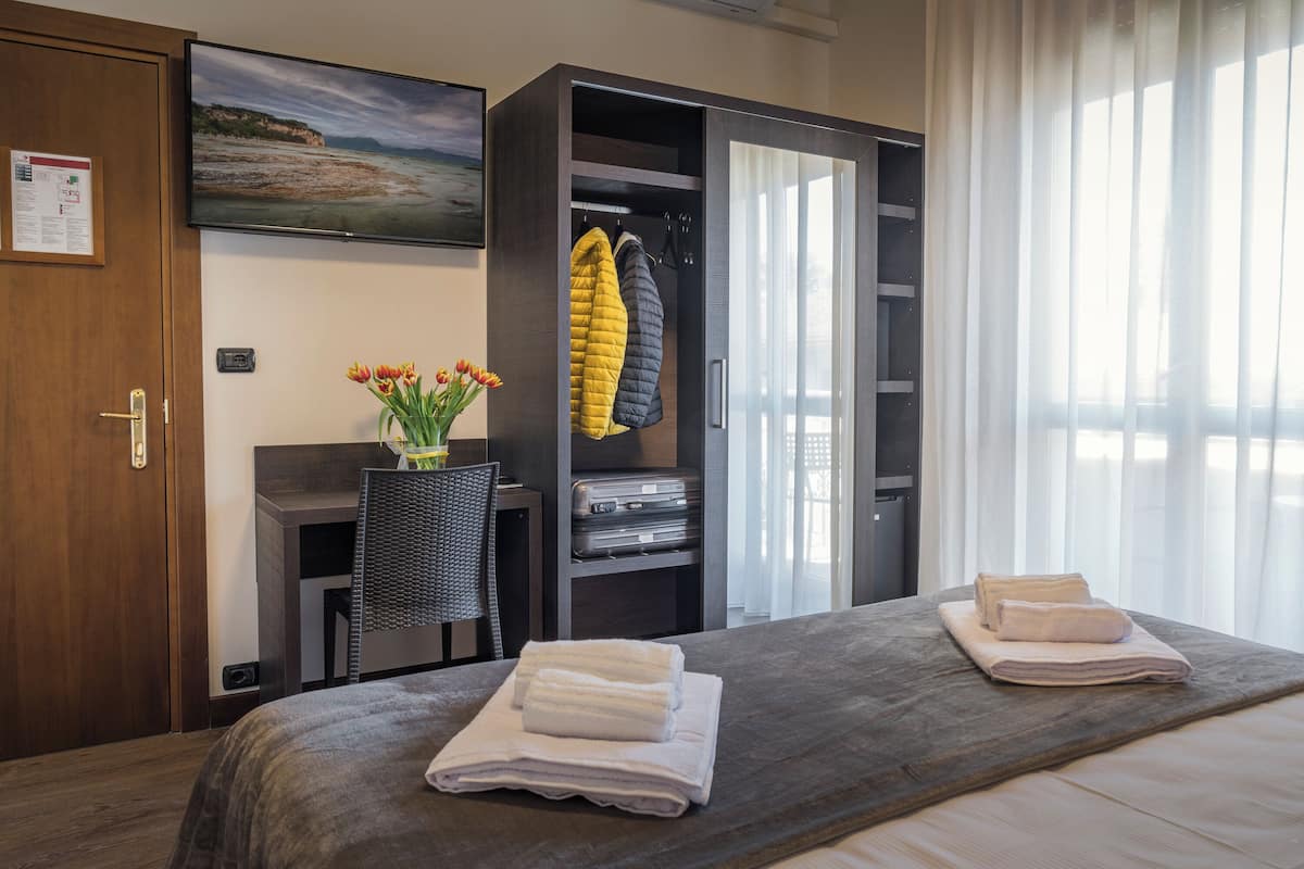 Quarto de hotel com cama, escrivaninha, guarda-roupa, televisão e janela com cortinas.