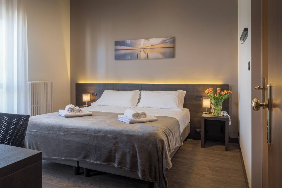Quarto de hotel com uma cama, abajures, uma pequena mesa com um vaso e um quadro na parede.
