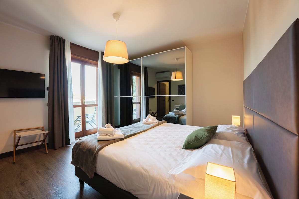 Quarto de hotel com cama grande, abajur, televisão de tela plana, espelho e janela com cortinas.