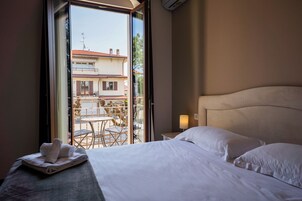 Una camera d'albergo con un letto, un comodino con una lampada, vista su un balcone esterno e un tavolo con sedie.