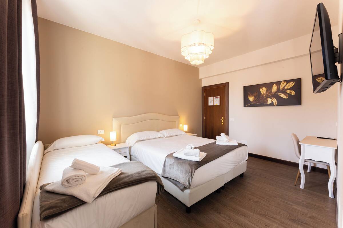 Quarto de hotel com duas camas, uma escrivaninha, uma cadeira, uma televisão e uma obra de arte na parede.