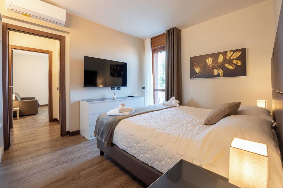 Quarto de hotel moderno com uma cama grande, uma TV de tela plana, uma escrivaninha com cadeira e uma janela com cortinas.