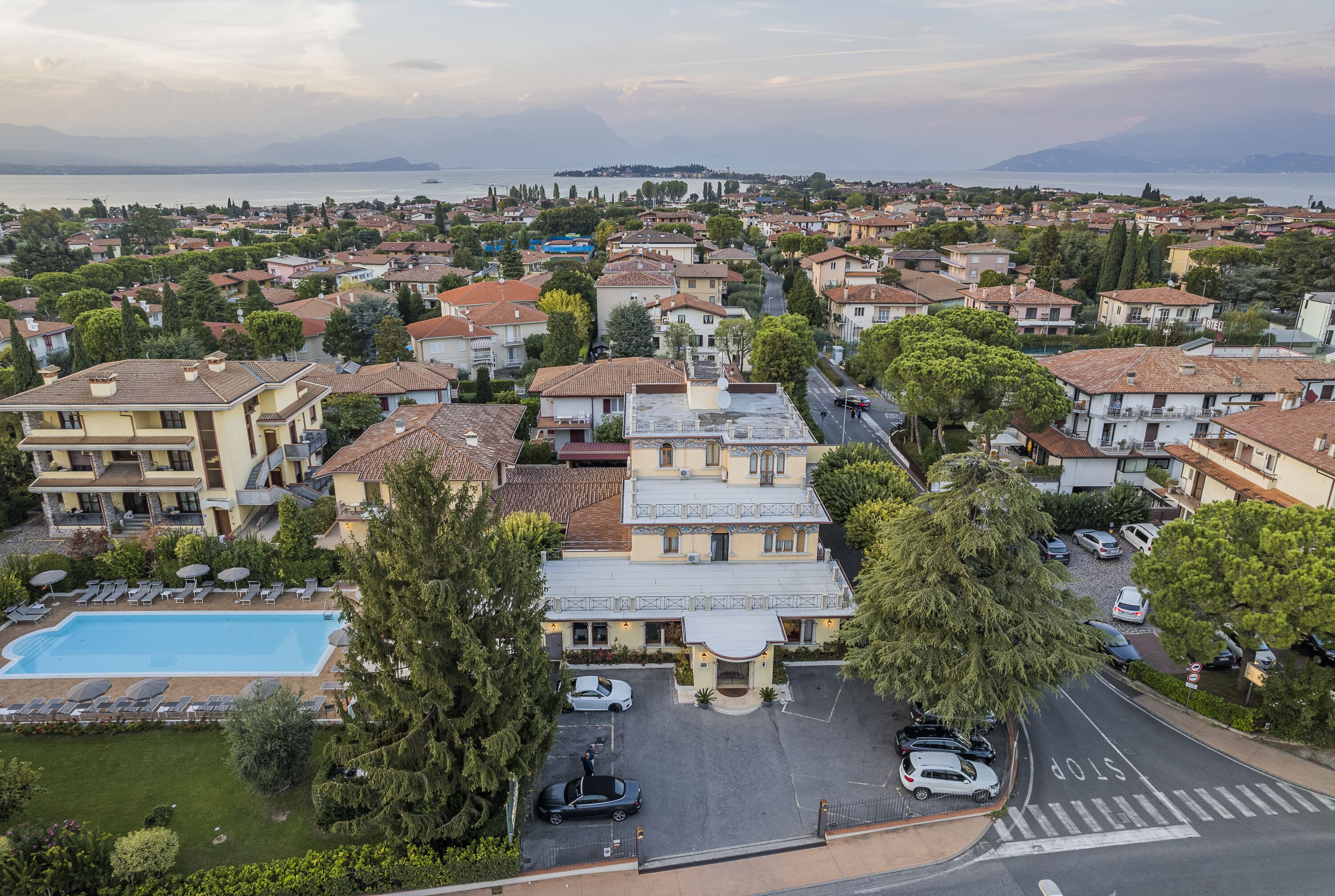 A residential area with a large swimming pool, multiple cars parked, and a view of the sea in the background.