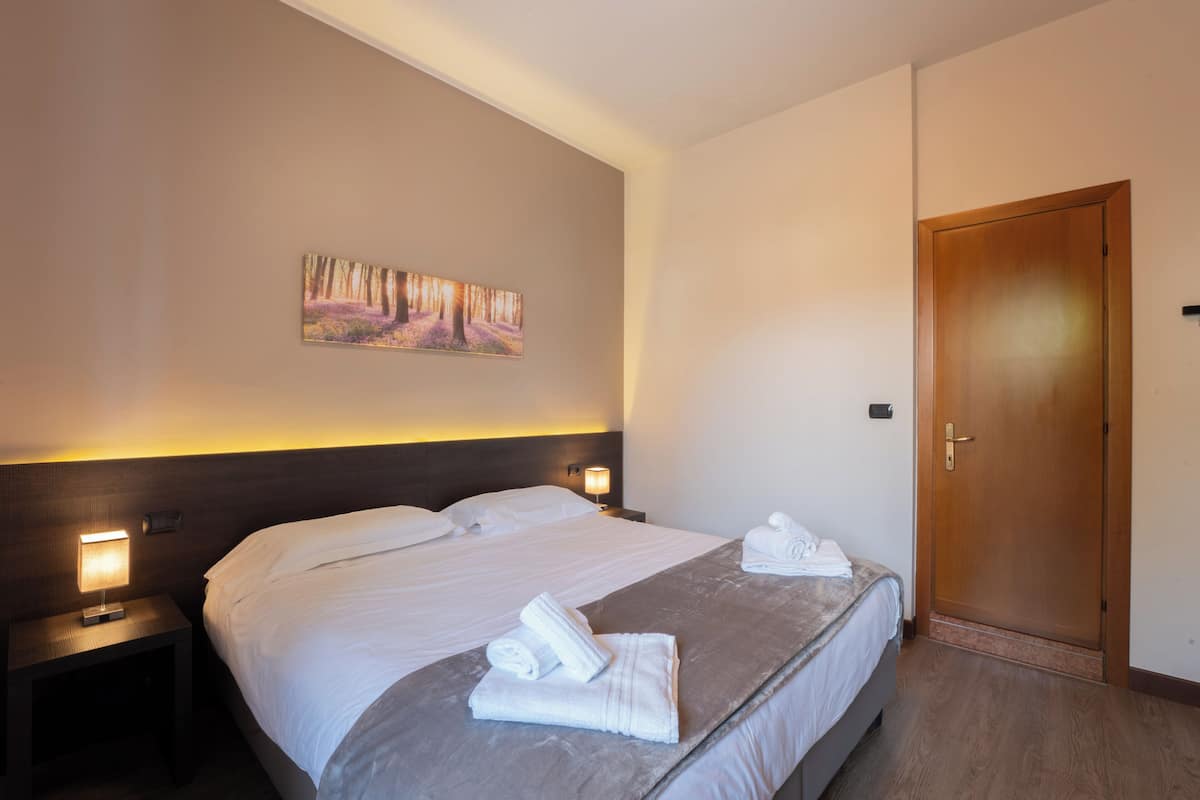 Quarto de hotel com cama, duas luminárias de cabeceira, um quadro na parede e uma porta de madeira.