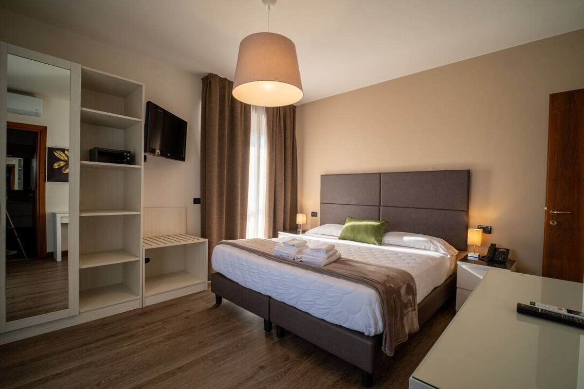 Quarto de hotel com cama, mesas de cabeceira, televisão, guarda-roupa e uma janela grande com cortinas.