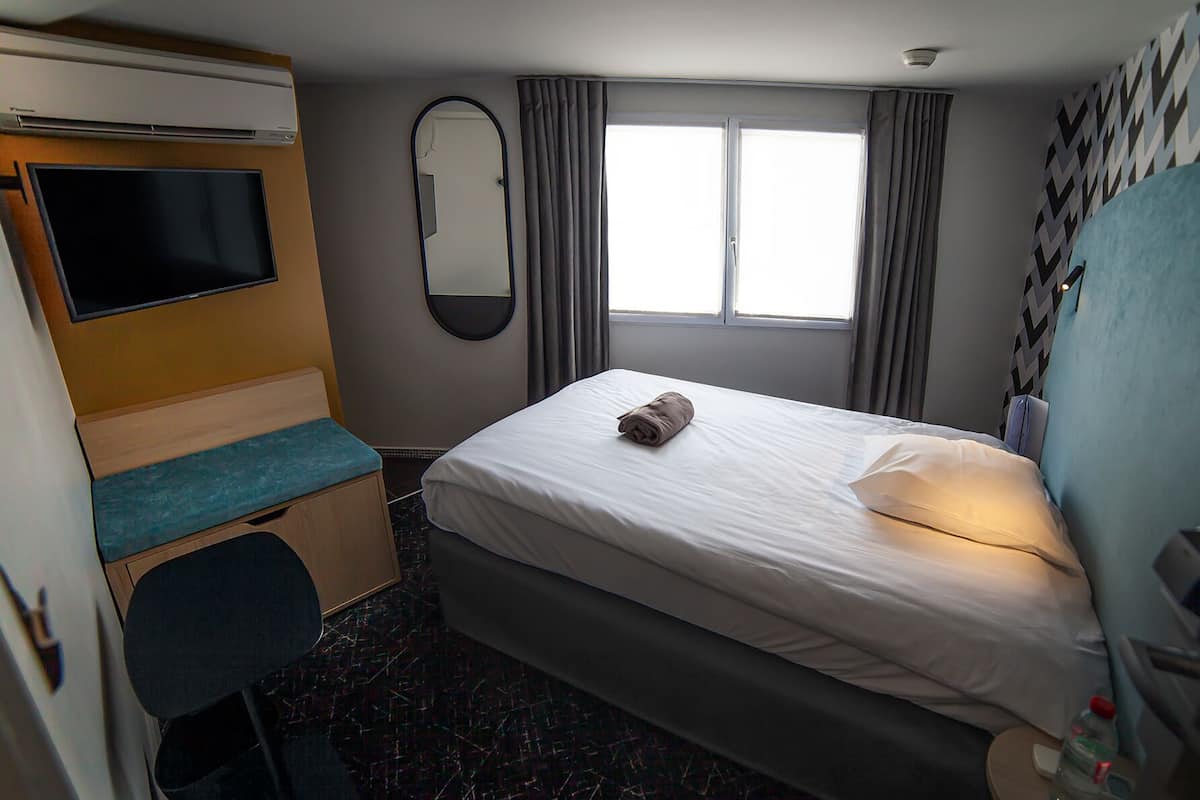 A hotel room with a bed, a desk with a chair, a television, a mirror, and a window with curtains.
