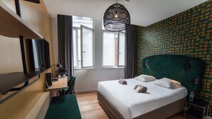 A hotel room with a large bed, a desk with a chair, a television, a window with curtains, and a patterned wall. - Hôtel Kanaï (Lille)