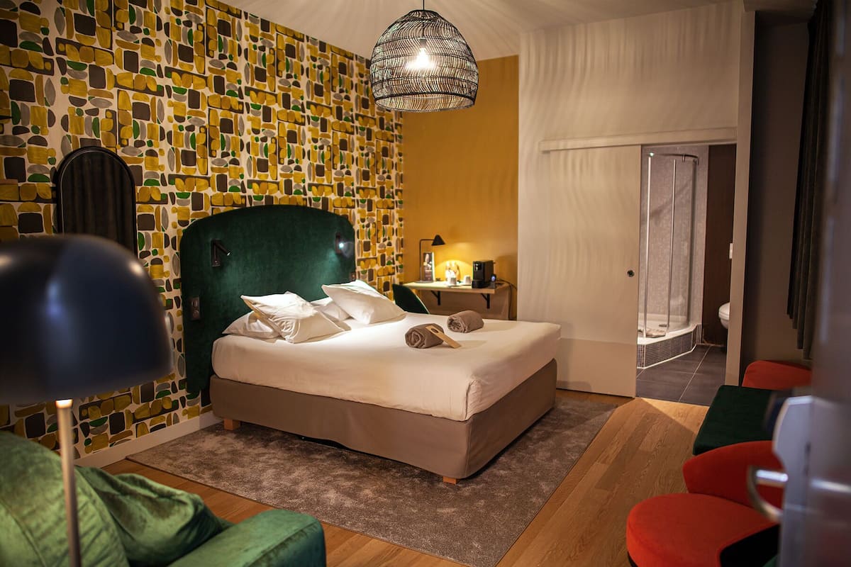 A modern hotel room with a patterned wall, a green upholstered headboard, a bed with white linens, a desk with a lamp, and a shower area.