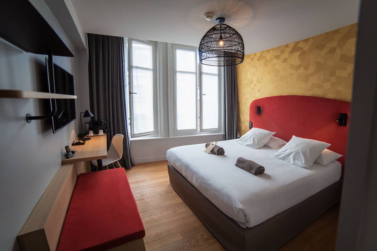 A hotel room with a bed, a desk, a chair, a TV, and a window with curtains.