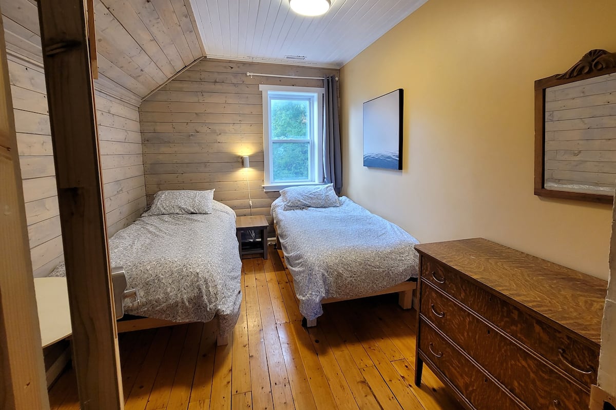 A small, cozy room with two beds, a wooden dresser, a nightstand, and a window with a view of trees.