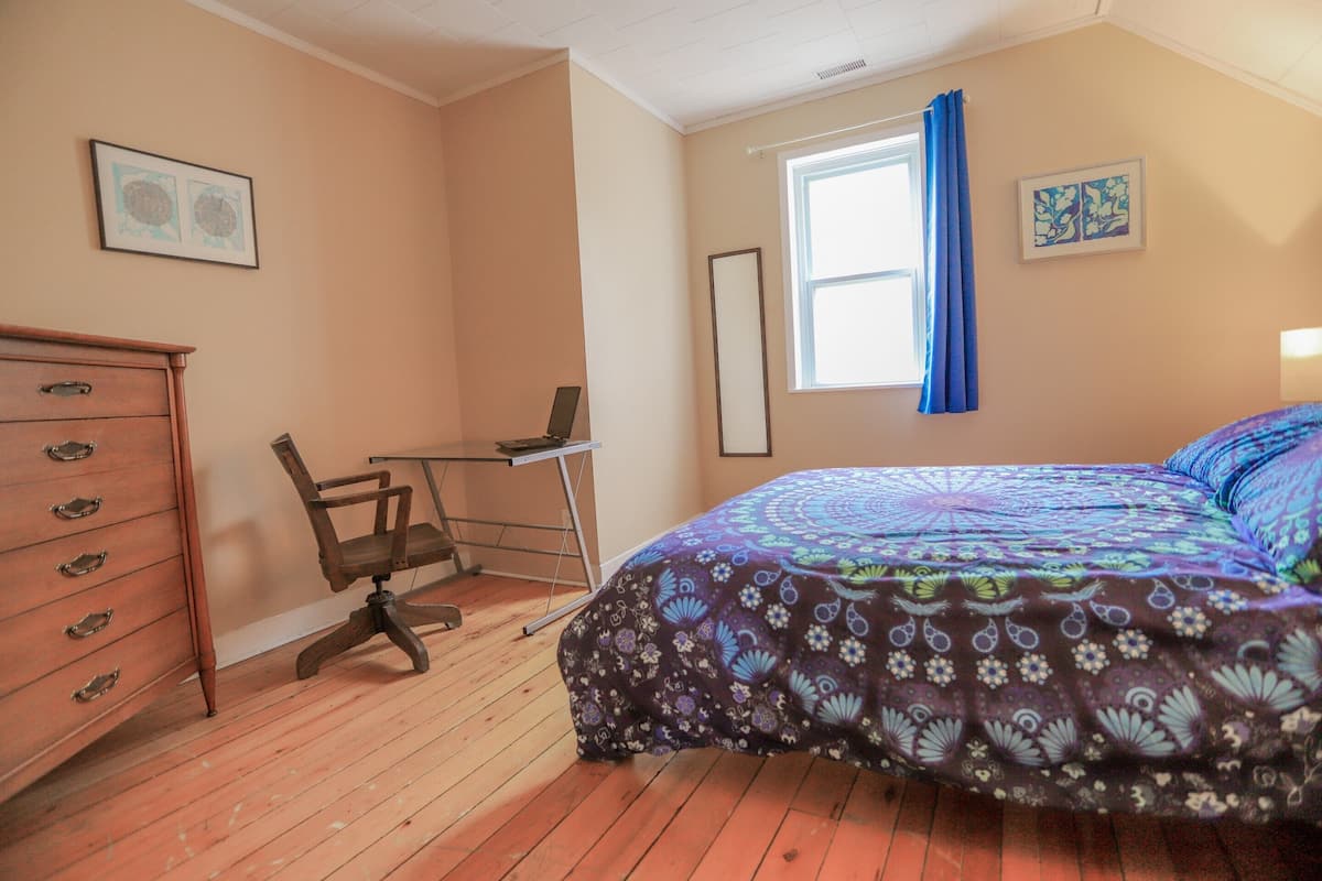 A bedroom with a wooden floor, a bed with a patterned bedspread, a desk with a chair, a dresser, and two framed artworks on the walls.