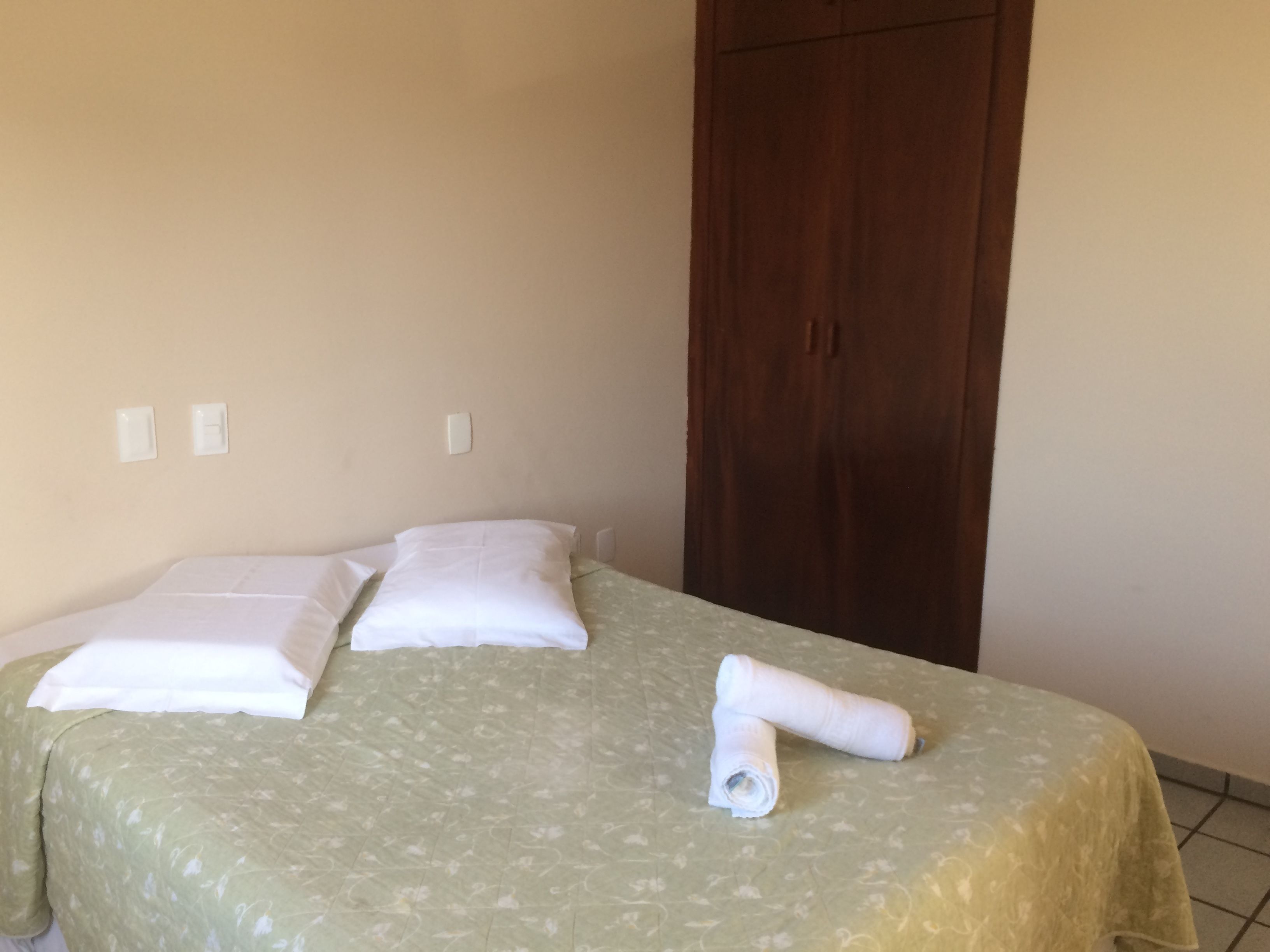 A single bed with a floral bedspread, two pillows, and a rolled towel on a plain wall with a wooden wardrobe.