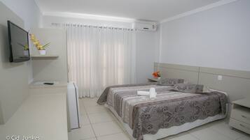 Quarto de hotel com cama, televisão, mesa pequena, planta e janela com cortinas.