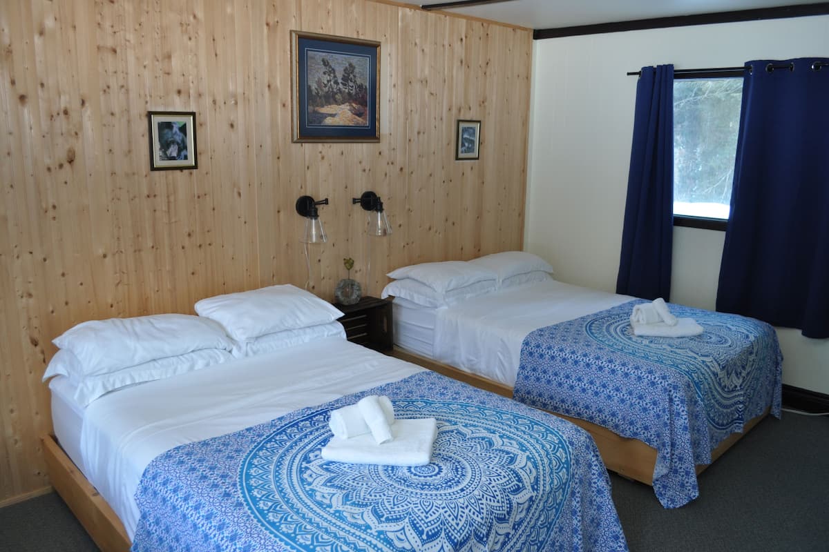 A hotel room with two single beds, wooden panel walls, framed pictures, a window with curtains, and a nightstand with lamps.