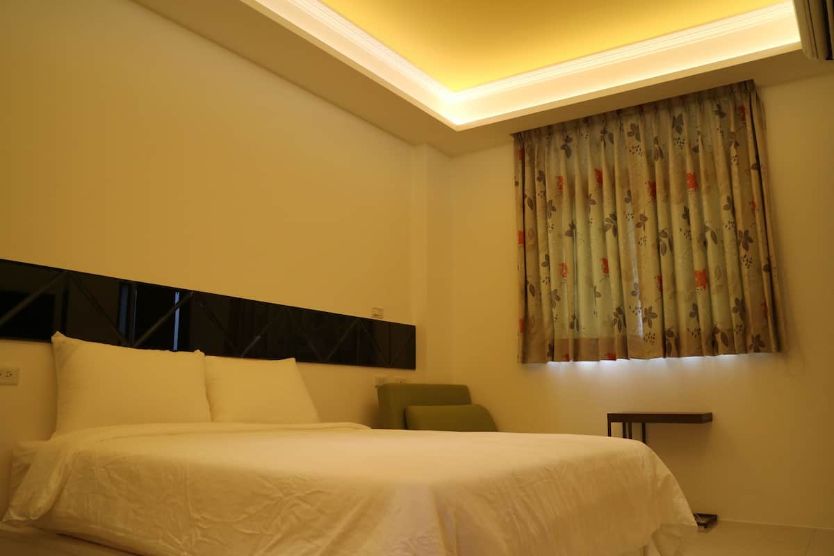 A hotel room with a bed, a headboard, a window with curtains, and a small table.