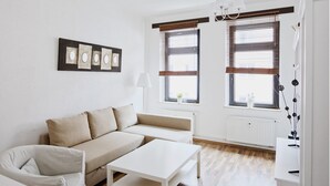 A modern living room with a beige sofa, white armchair, and a white coffee table. There are two windows with blinds, a white radiator, and a wall-mounted light fixture. - City Park Apartment 23 (Leipzig)