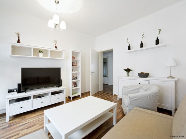 A modern living room with a flat-screen TV, white furniture, and a bookshelf.
