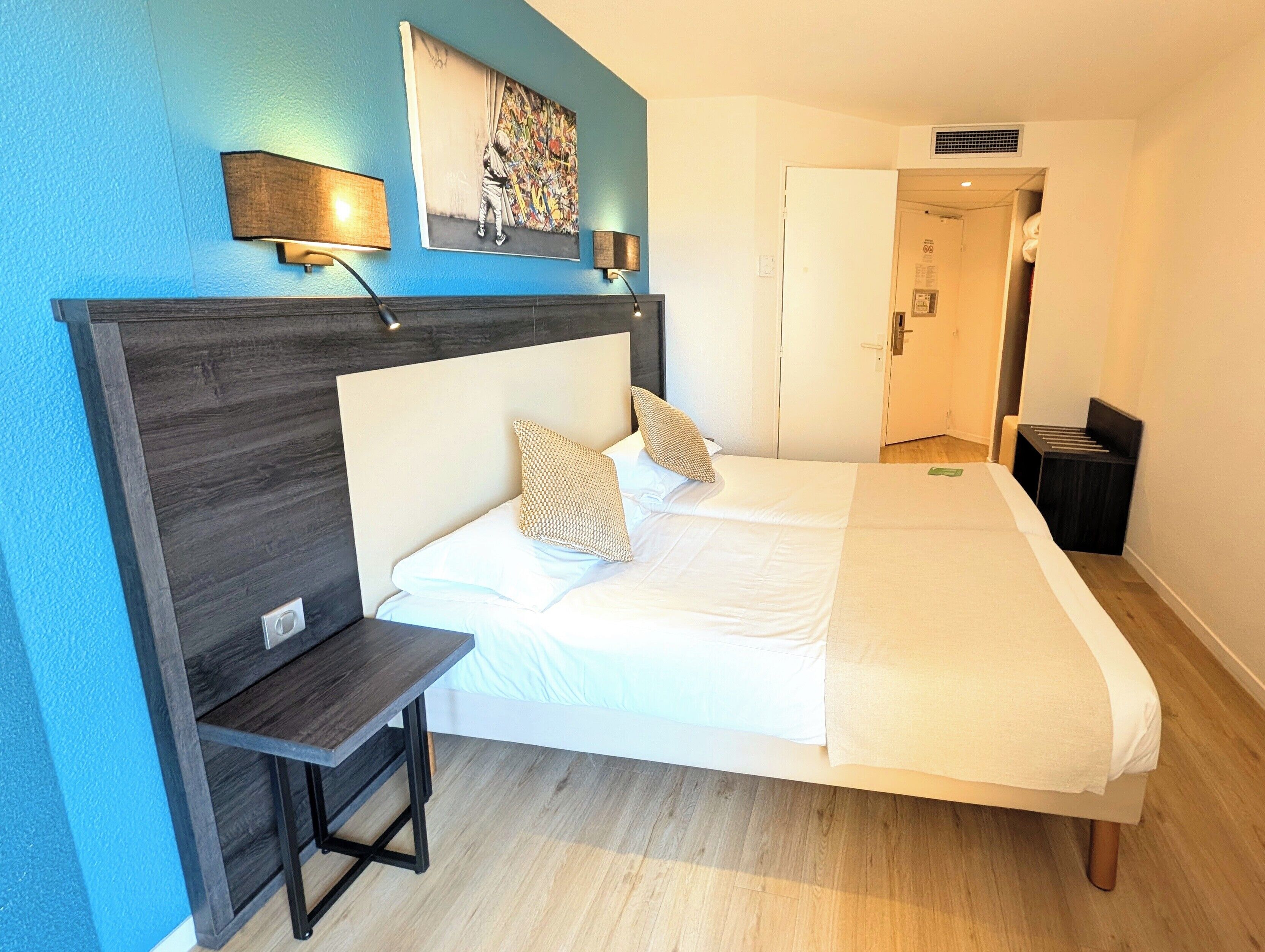 Une chambre à coucher comprenant un lit, une table de chevet, une lampe, des œuvres d’art et une porte donnant sur une salle de bain.