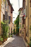 A narrow cobblestone street lined with buildings, some with shutters and plants. A sign for a hair salon is visible.