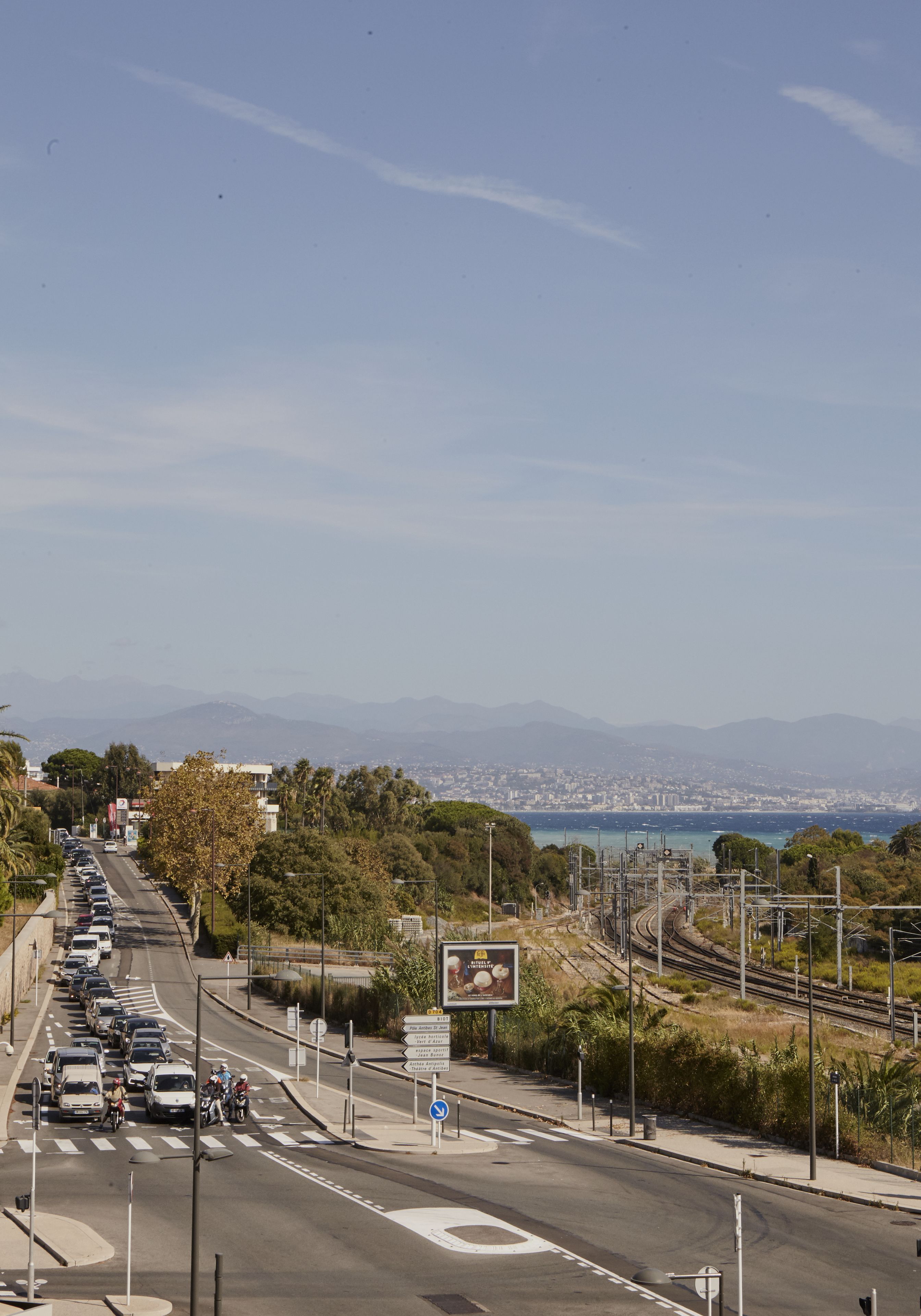 Une longue portion de route où l’on aperçoit des voitures et des motos, un passage piéton, et une vue dégagée sur des montagnes lointaines et une étendue d’eau.