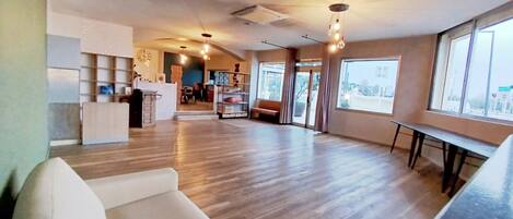 A spacious living area with a wooden floor, a white sofa, a dining table, and a kitchen area with a clock on the wall.