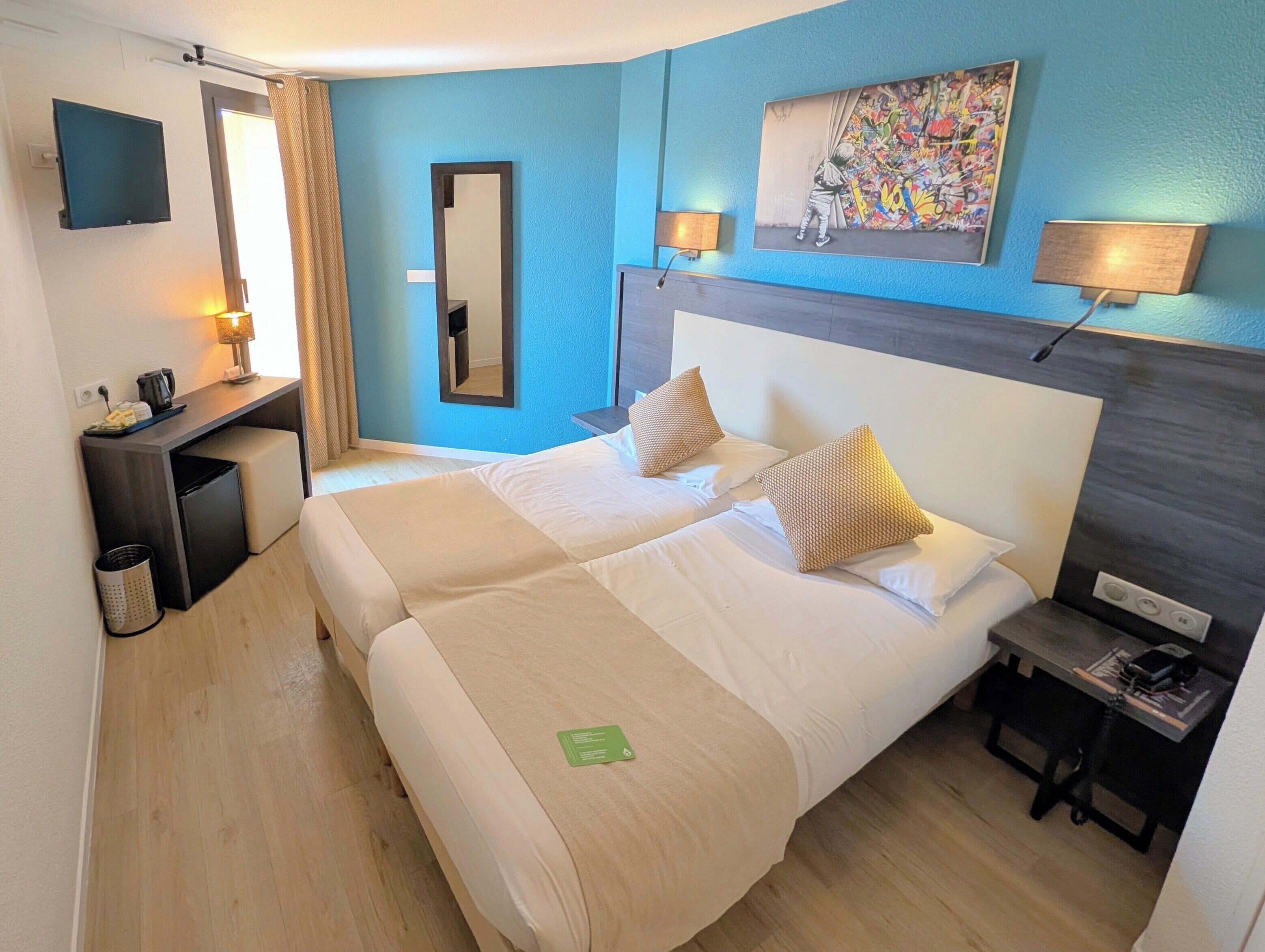 Habitación de hotel con dos camas, un escritorio, un espejo, un cuadro y una pequeña mesa con lámpara.