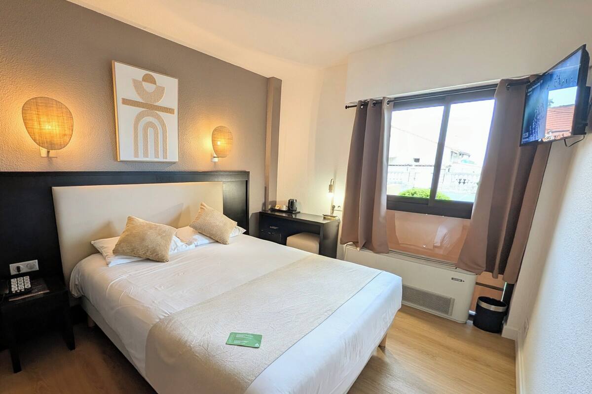 Habitación de hotel con una cama grande, mesitas de noche, televisión montada en la pared y una ventana con cortinas.