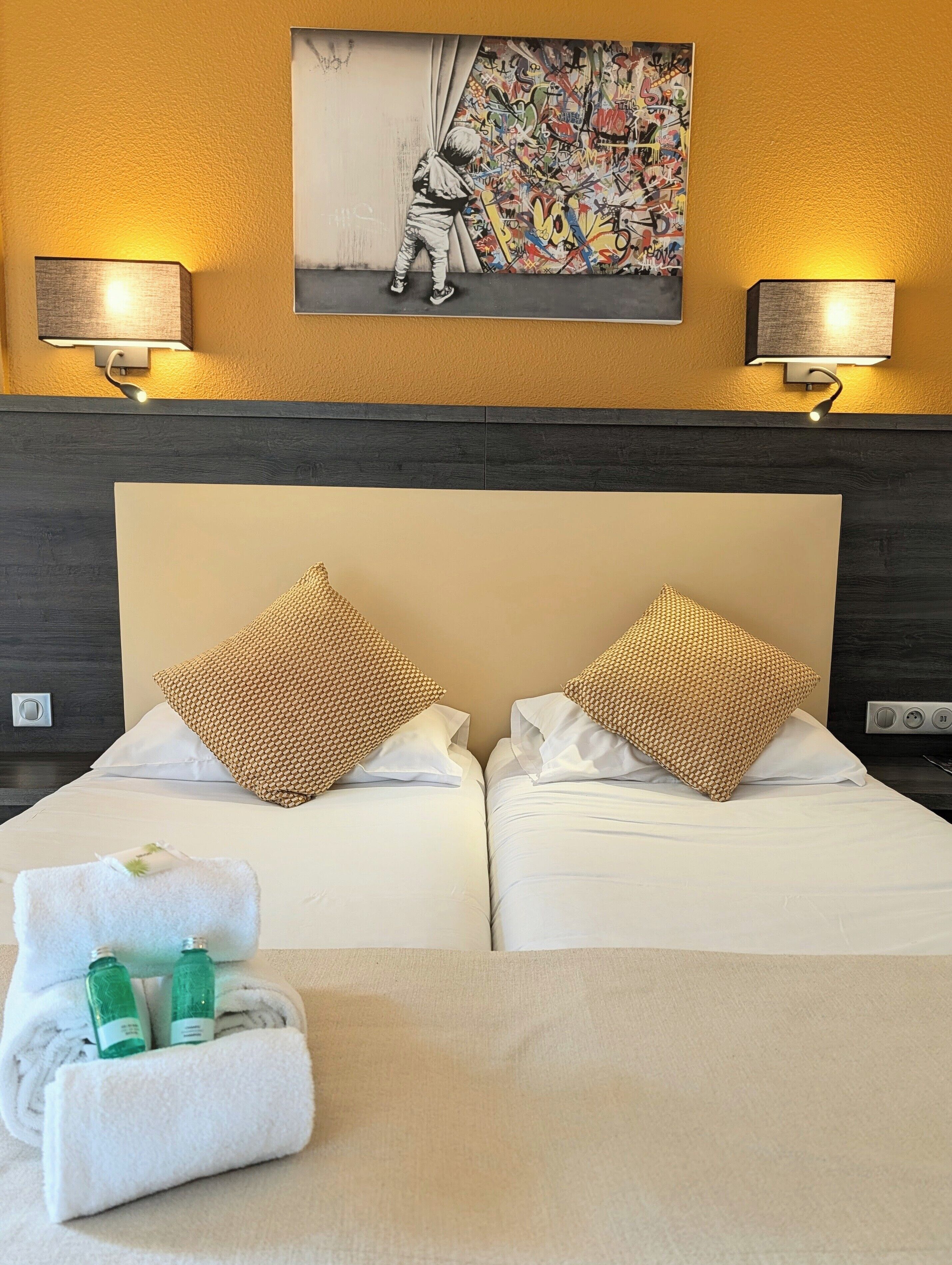 A hotel room with two beds, a wall-mounted artwork, and two wall-mounted lamps.