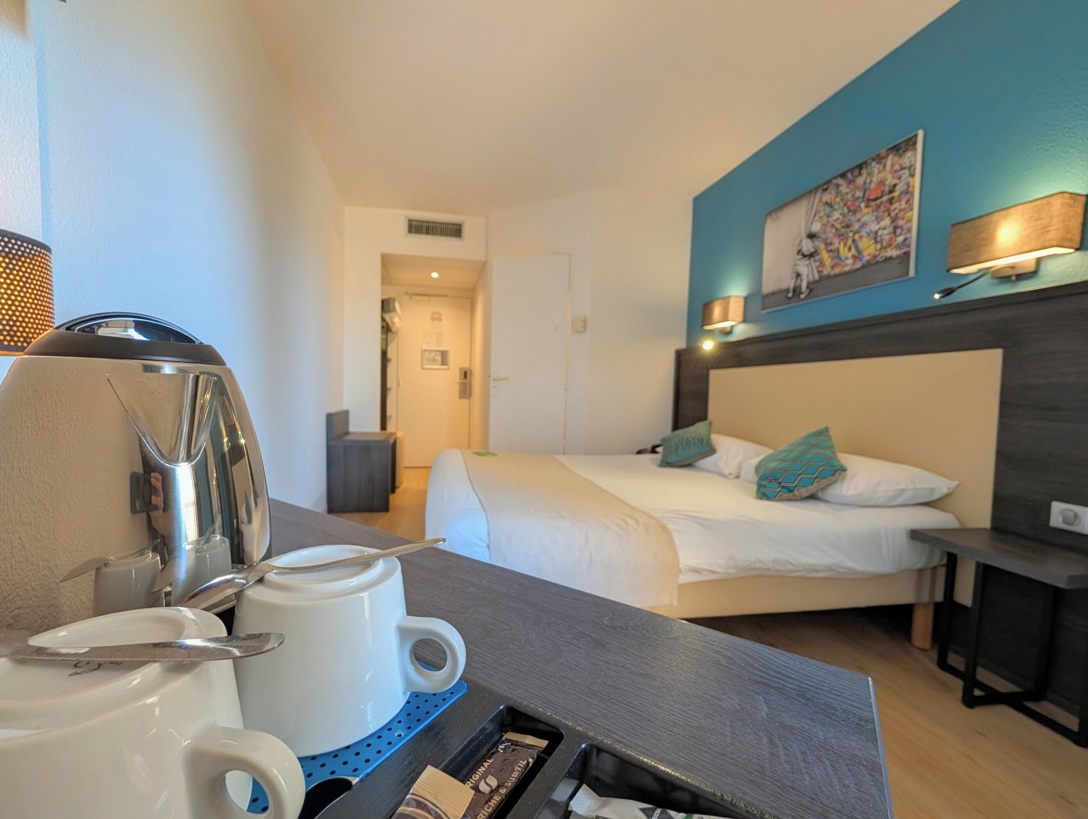 Quarto de hotel com cama, mesa de cabeceira, cafeteira e um quadro na parede.
