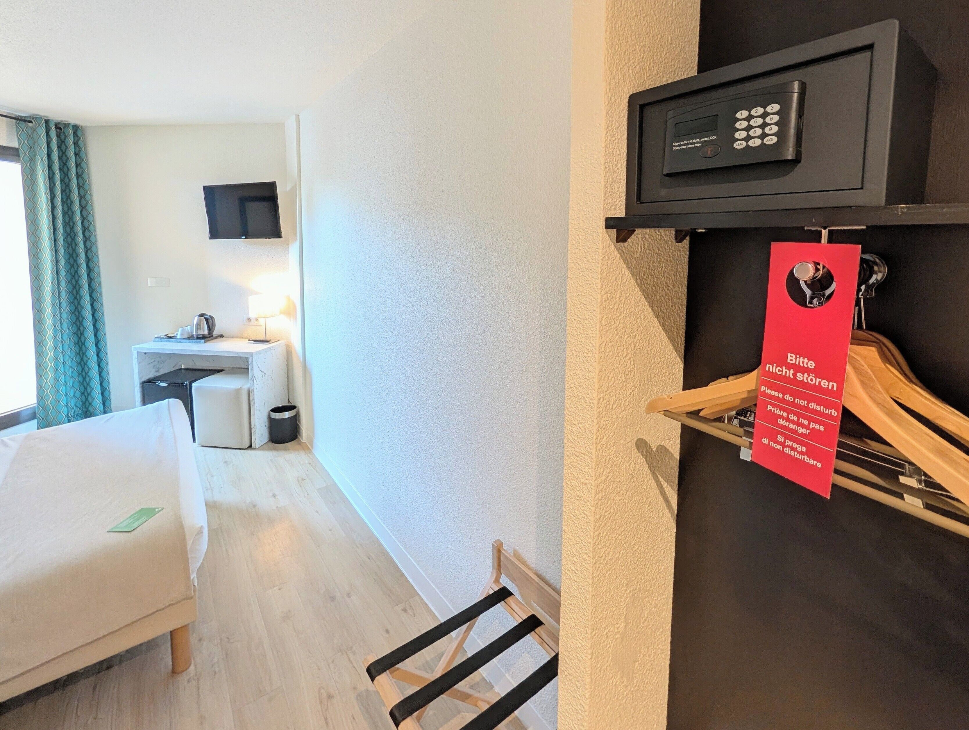 A hotel room with a bed, a desk with a lamp, a TV, a coat rack, and a red sign with a message.
