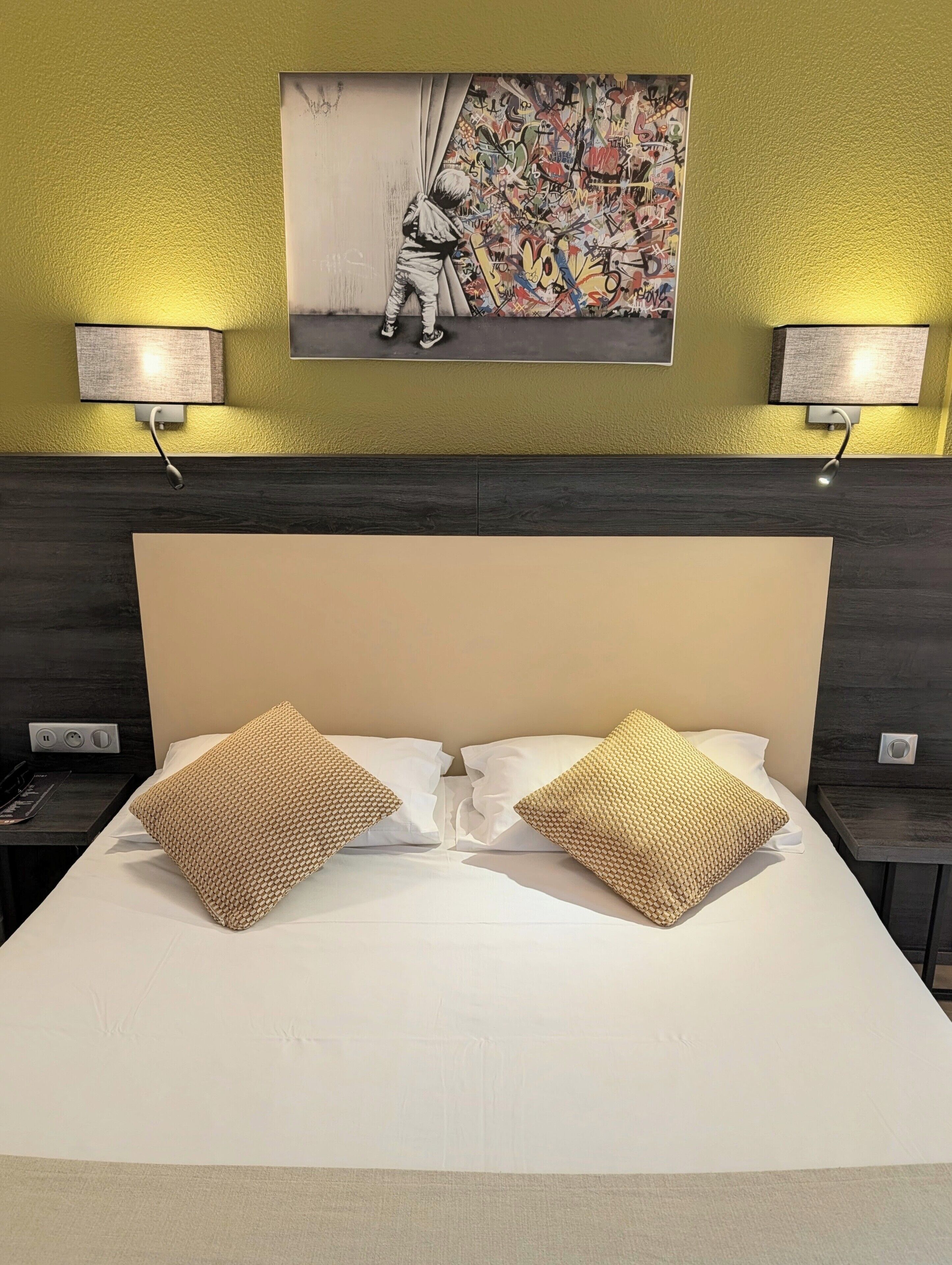 A hotel room with a large bed, two bedside lamps, a wall-mounted artwork, and a dark wood headboard.