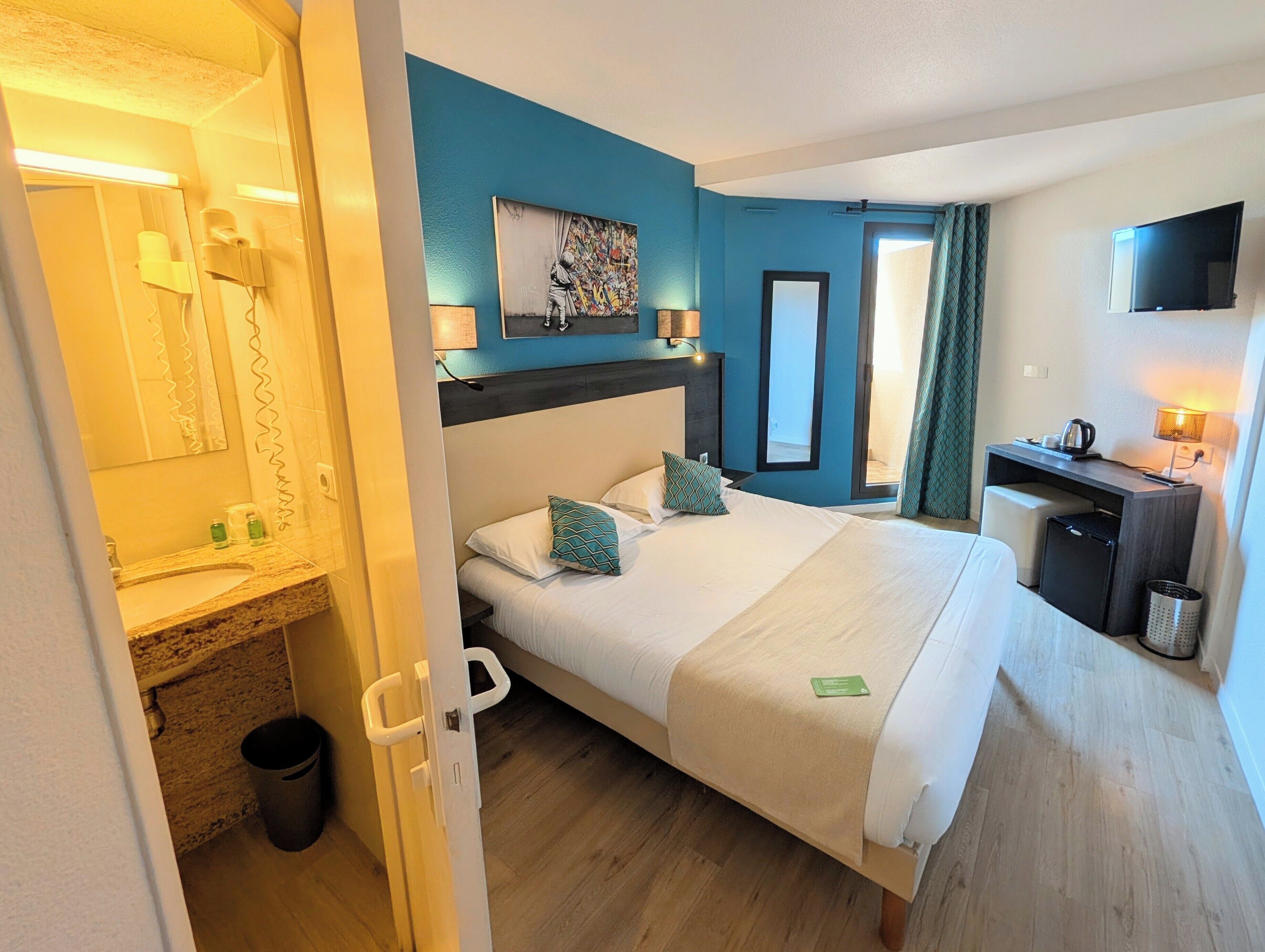 A hotel room with a bed, a desk, a TV, a bathroom, and a painting on the wall.