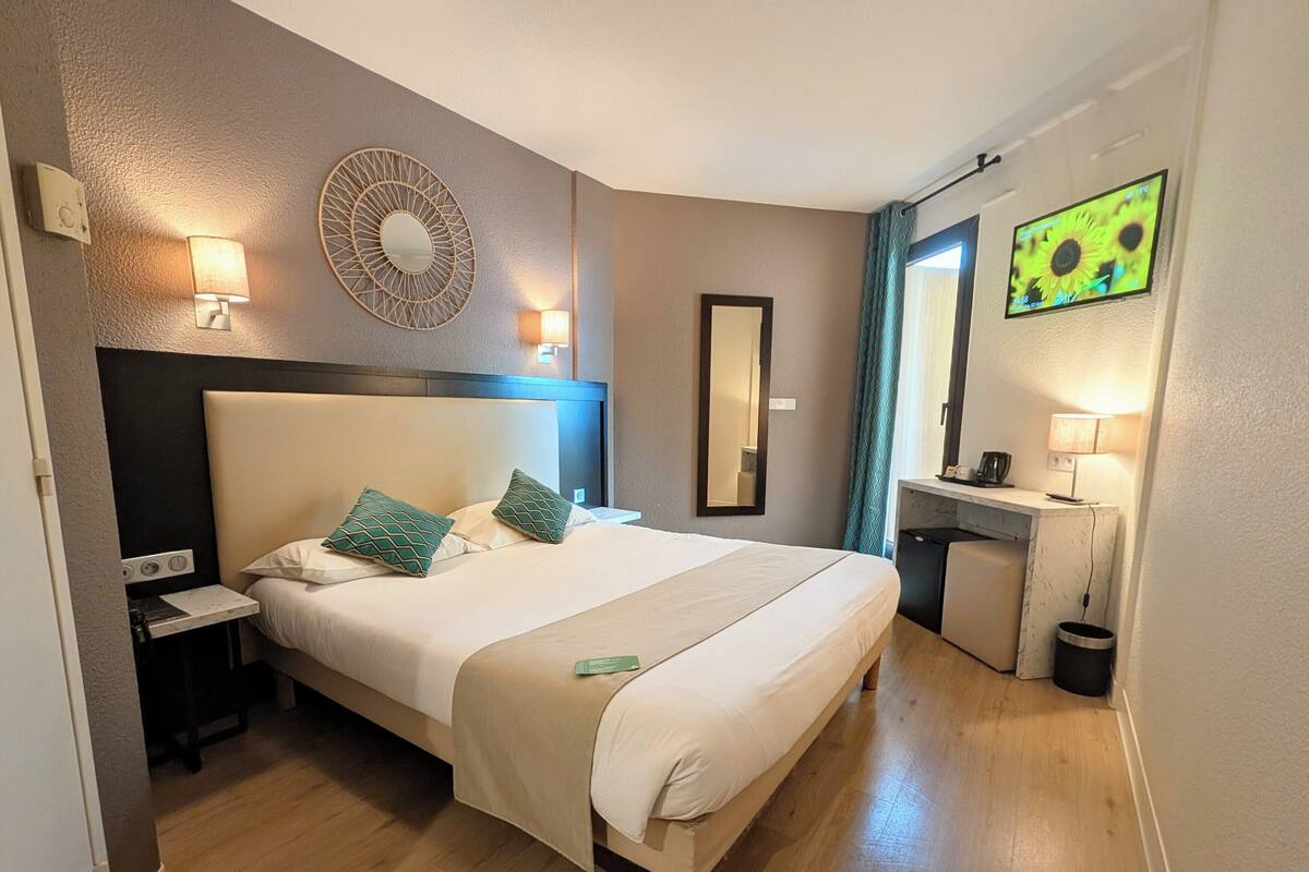 Una habitación de hotel con cama, mesitas de noche, un escritorio, un espejo y una televisión.