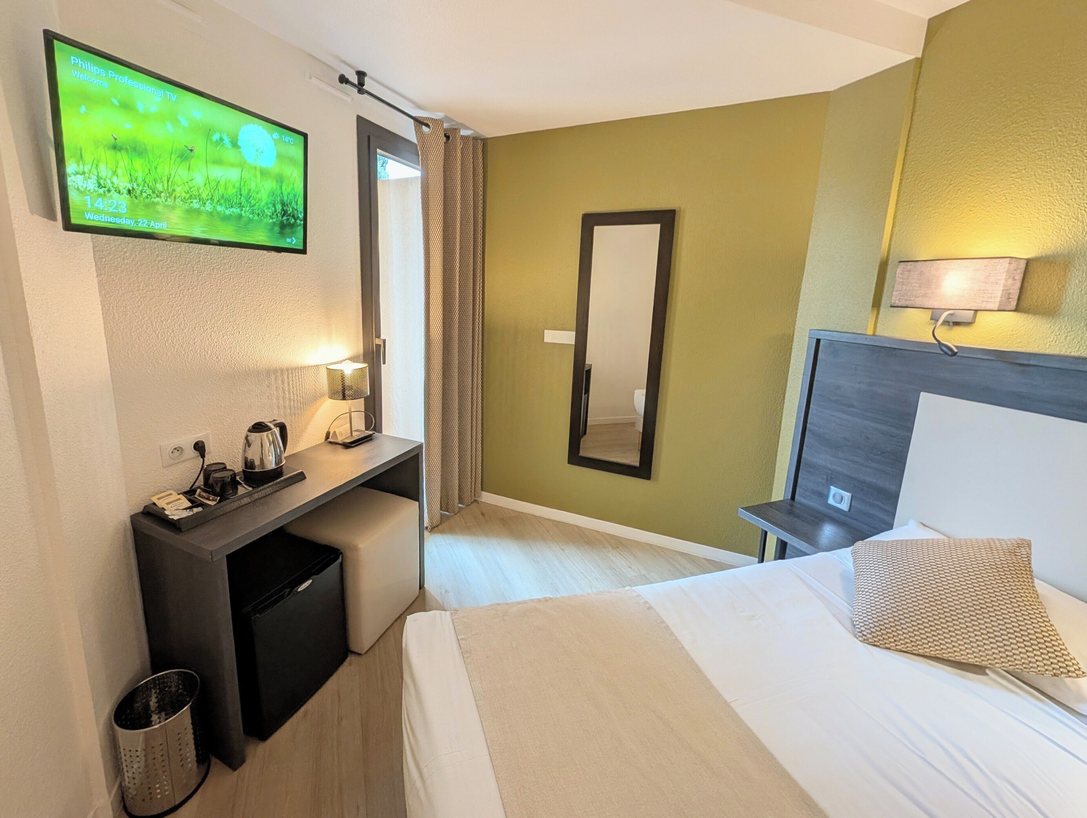 Una habitació d'hotel amb un llit, una taula de nit amb una làmpada, un televisor, un mirall i un petit safataire.