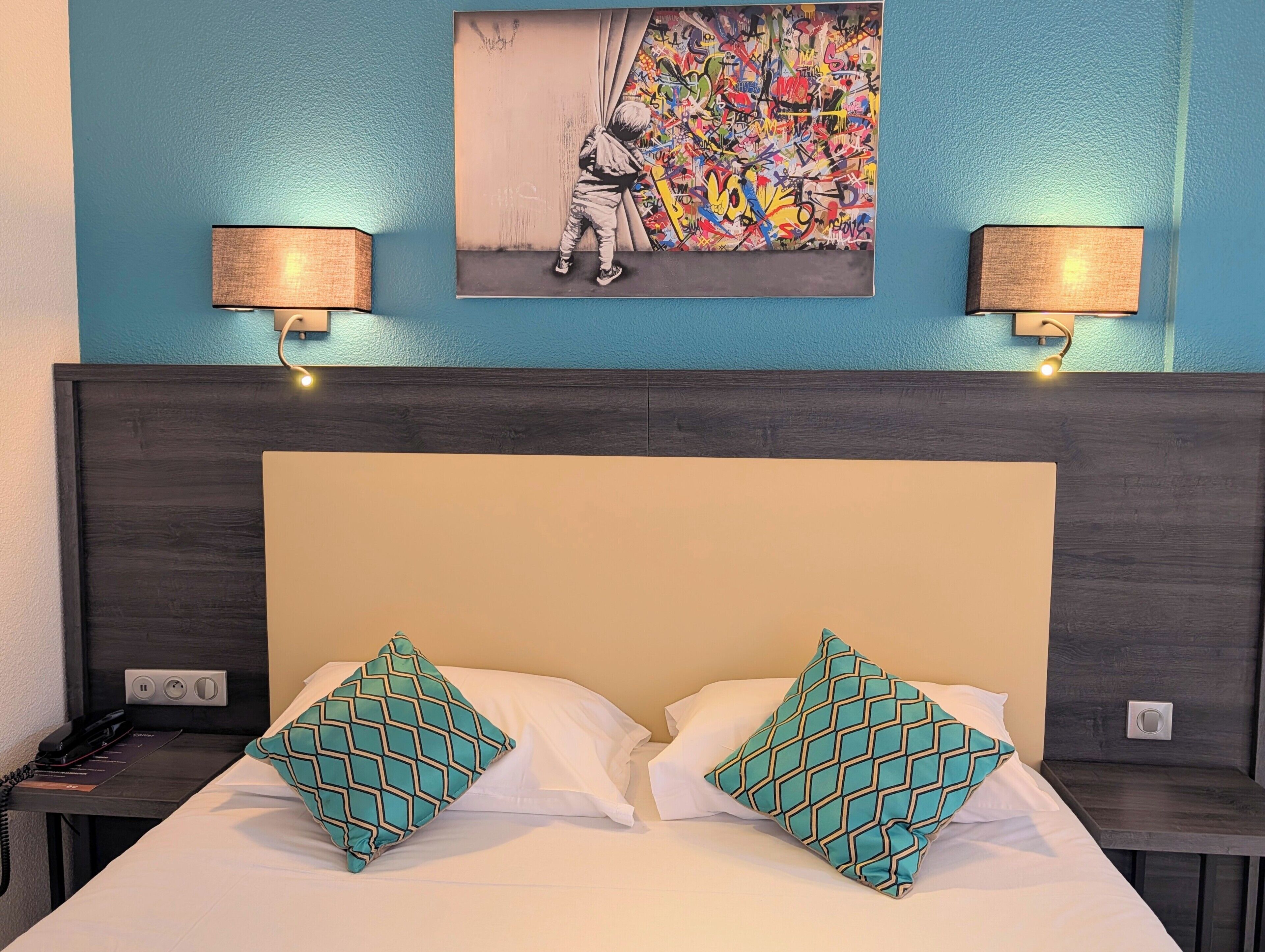 Une chambre d’hôtel avec un lit, deux coussins à motifs, une œuvre d’art accrochée au mur et deux lampes murales.
