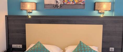 A hotel room with a bed, two patterned pillows, a wall-mounted artwork, and two wall lamps.